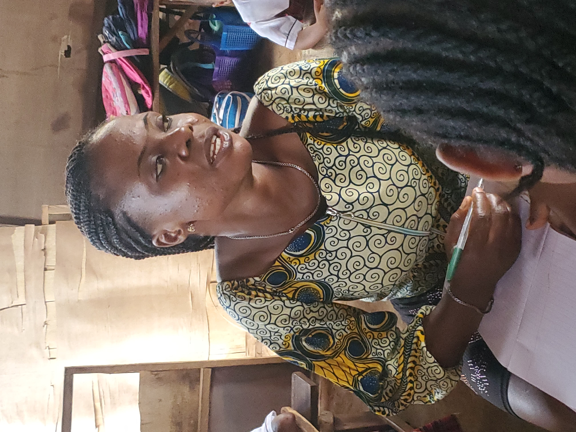 In Ukwu-Abwa, Nigeria, an African woman with braided hair is seen in a room. She wears a dress with a cream-colored background and a spiral pattern in dark blue and yellow. She holds a pen and appears to be writing on a sheet of paper. Another person with dark hair is partially visible in the foreground. In the background, there are bags and shelves. The room appears modestly furnished.