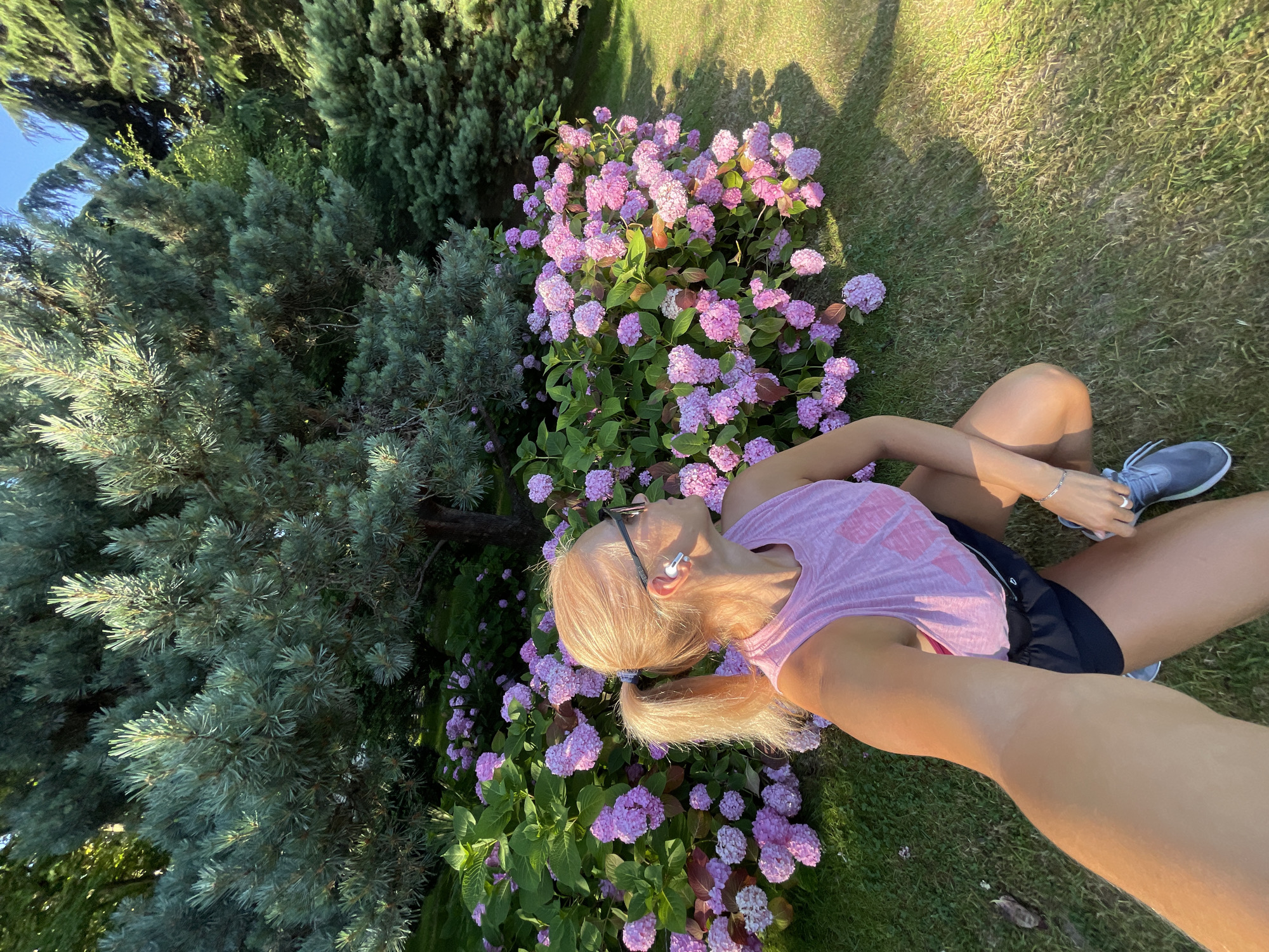 A woman with blonde hair in a ponytail, wearing a pink tank top, black shorts, and gray sneakers, is captured in a selfie taken outdoors. She is squatting on a grassy lawn in front of a dense bush of pink hydrangeas and a large, blue-green conifer tree. The scene is illuminated by warm, late afternoon sunlight, casting long shadows on the grass. The background consists of more trees and foliage, suggesting a park or garden setting. The location is identified as Sochi, Russia. The woman is wearing white earbuds and sunglasses. She appears to be looking to her right, away from the camera.