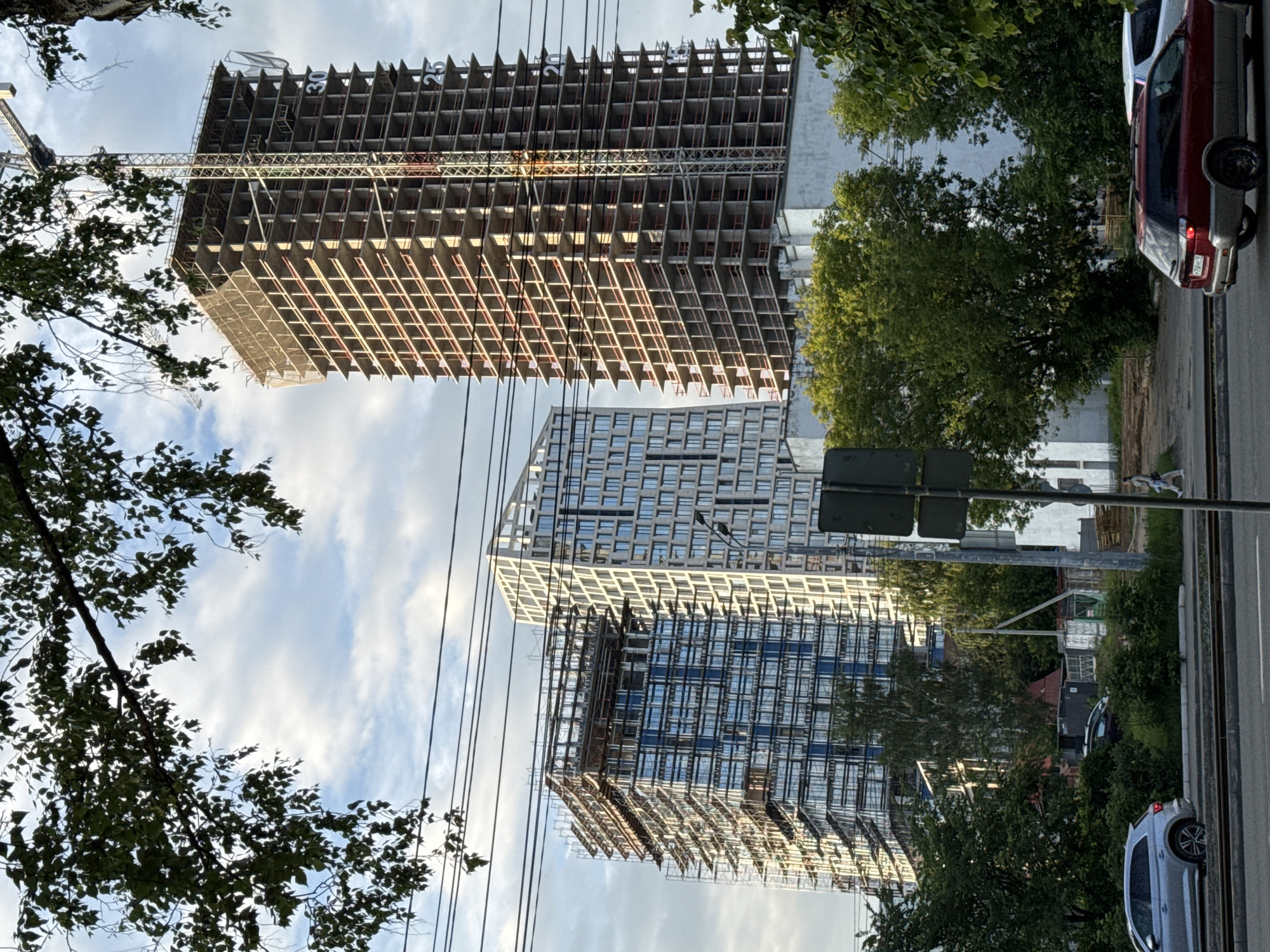 In Izhevsk, Russia, a construction site features two high-rise buildings under development, situated amidst a partly cloudy sky. On the right, a taller, unfinished concrete skyscraper is visible, equipped with a construction crane. The building's floors are marked with numbers, including '30' and '25'. To the left, a shorter, more completed building displays a façade of blue windows and is partially covered in scaffolding. In the foreground, a multi-lane road runs horizontally, with cars, including a silver SUV and a red station wagon, navigating the lanes. Trees and overhead power lines are present in the scene, contributing to the urban environment.