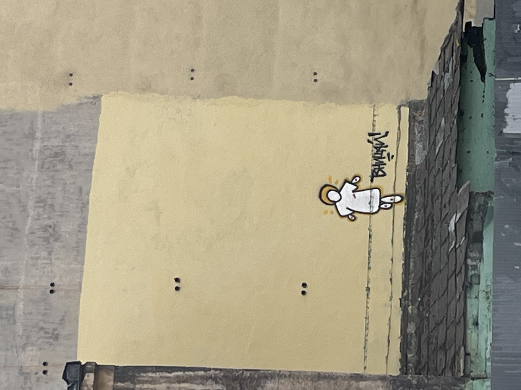 The image depicts a section of an exterior wall, likely in Saint Petersburg, Russia, based on the Cyrillic writing. The wall is painted in sections of gray and yellow. On the yellow section, there is a graffiti-style image of a white stick figure with a yellow outline, lying horizontally. To the right of the figure, there is some stylized Cyrillic lettering. To the right of the painted wall, there is dilapidated roofing. Above the gray and yellow painted section of the wall, there are two sets of two small, dark dots in the lighter shade of yellow. There is an additional section of gray towards the bottom of the wall.