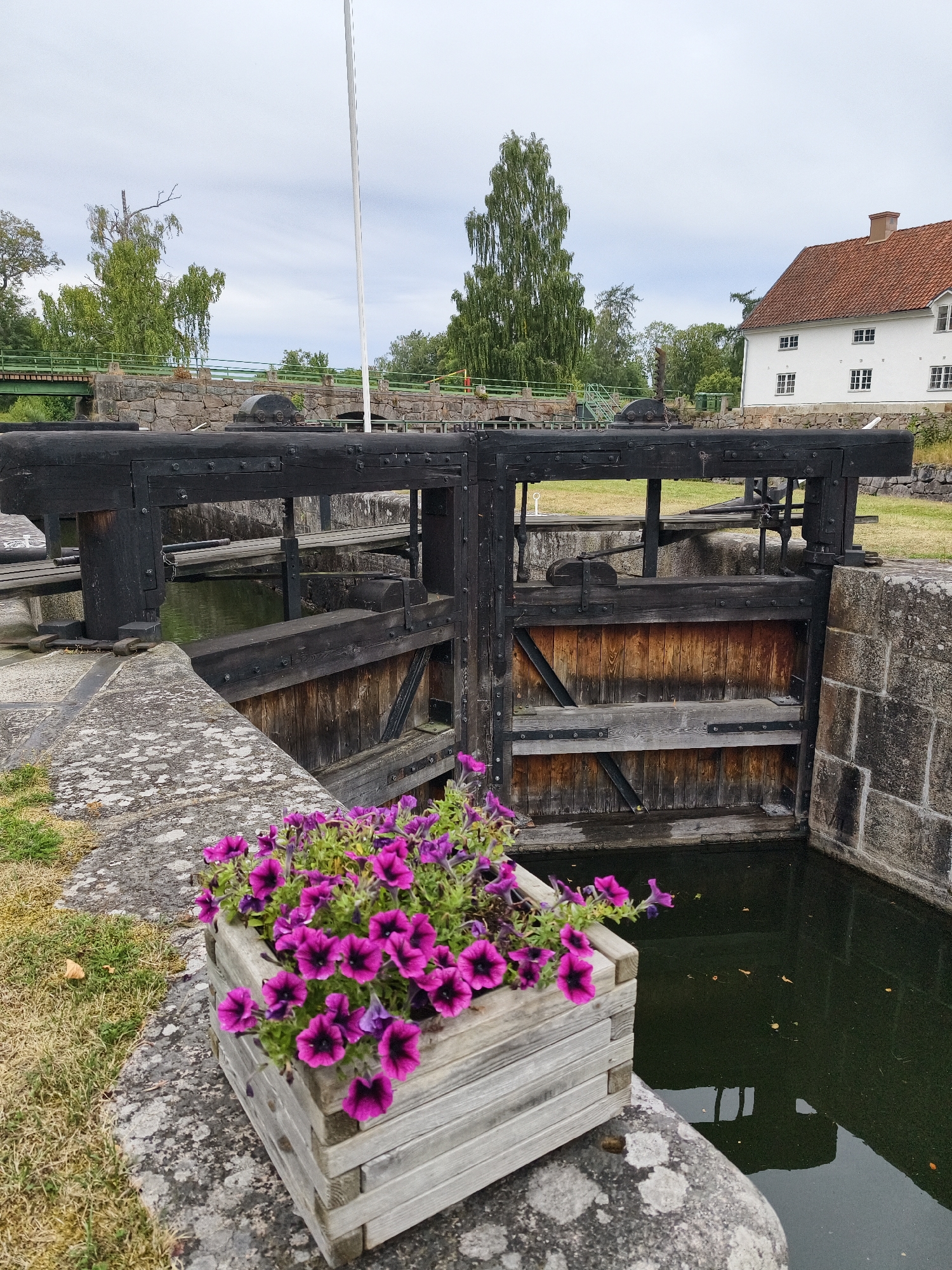 The image shows a canal lock in Brokind, Sweden. The lock is made of dark wood with metal reinforcements and is surrounded by stone walls. In the foreground, a wooden planter box filled with vibrant purple petunias sits on a stone ledge. The water in the lock is dark and still, reflecting the wooden gates and the overcast sky. Beyond the lock, a white building with a red roof and a green bridge can be seen. The overall scene depicts a peaceful, rural setting.