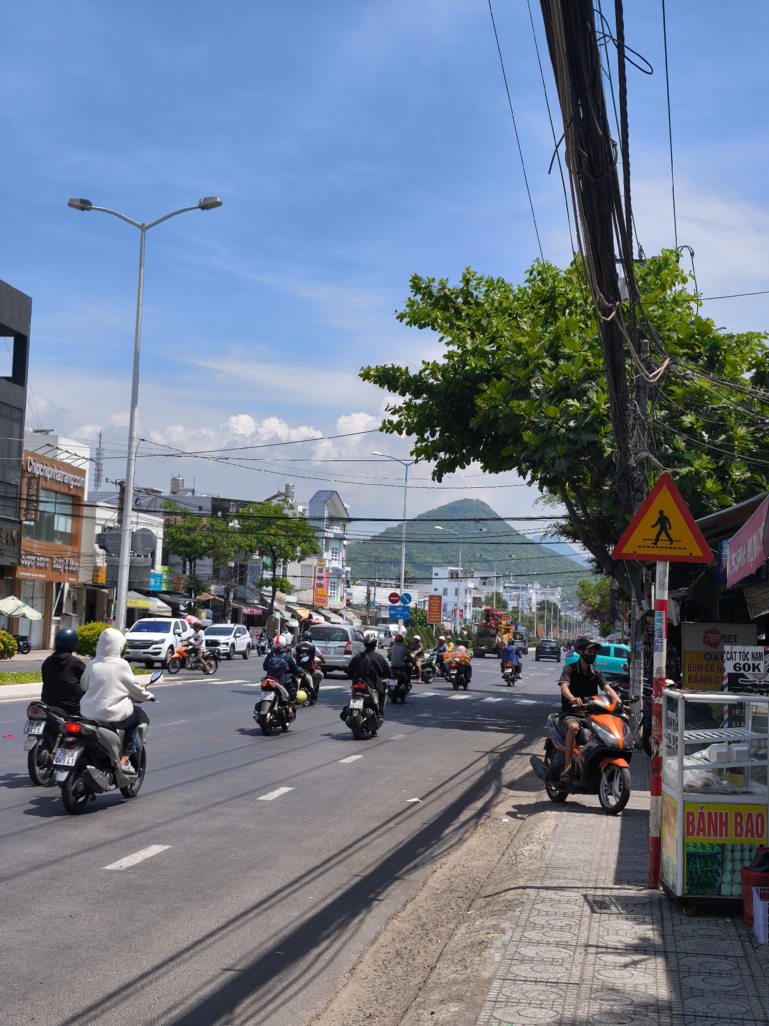 The image depicts a street scene in Bac Nha Trang Ward, Vietnam, on a bright, sunny day with a blue sky dotted with clouds. A multi-lane asphalt road is the central focus, with several motorbikes and a few cars navigating through traffic. The motorbikes are predominantly ridden by individuals wearing helmets, and some are carrying passengers. On the right side of the road, a pedestrian walkway with patterned tiles is visible, bordering a food stall with a display of various items, including what appears to be baked goods. A prominent yellow triangular sign with a pedestrian crossing symbol is posted on a pole. Buildings line the left side of the street, displaying signage in what appears to be Vietnamese. Lush green trees are present on both sides of the street and in the background, with a mountain visible in the distance. Overhead, a complex network of electrical wires hangs from poles, contributing to the urban streetscape. The overall impression is of a bustling street in a Vietnamese city.