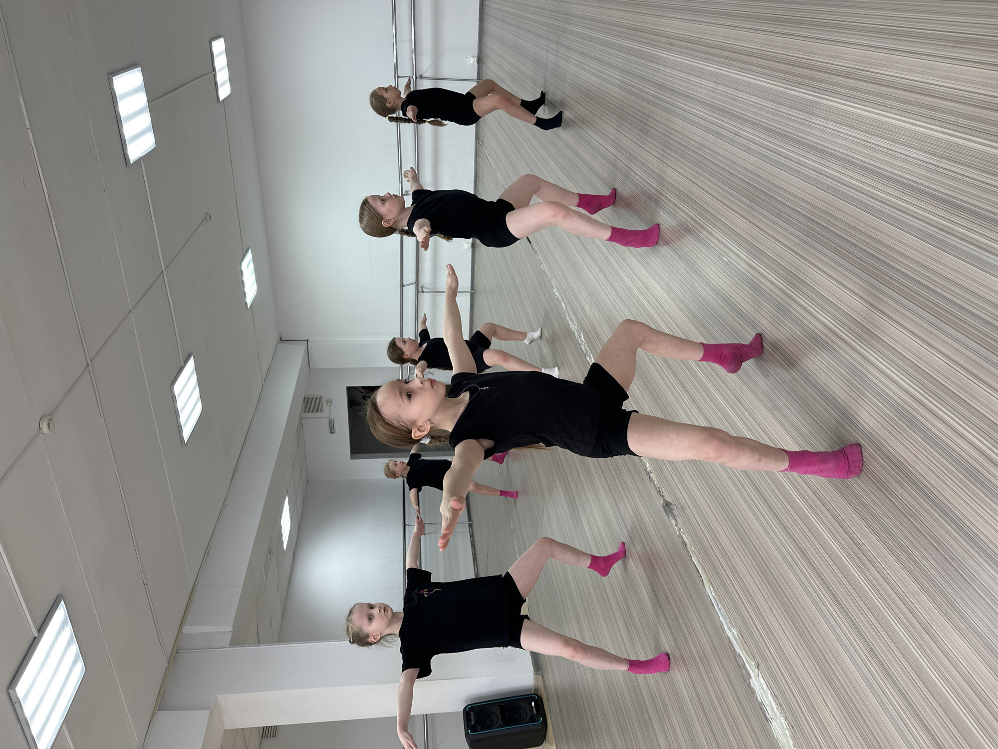 This image depicts a group of approximately seven to eight young girls engaged in a dance or exercise class within an indoor studio in Votkinsk, Russia. The studio features a light-colored wooden floor with distinct horizontal stripes, white walls, and rectangular fluorescent lights illuminating the space. A dance barre runs along one wall, reflected in a large mirror that also shows parts of the room.
The girls, appearing to be pre-teens, are uniformly dressed in black t-shirts and black shorts. Most are wearing bright pink socks, which stand out against their dark attire, though one or two wear black or white socks. Their hair is neatly tied back, with some styled in braids. They are performing a floor-based exercise, lying on their backs with their arms extended horizontally to the sides and legs bent or slightly raised, demonstrating a pose often used for core strength or flexibility training. Their faces convey concentration and discipline.
A black portable speaker with the "SONY" brand visible is positioned on the floor in the foreground. The photograph is taken from a low, tilted angle, creating a dynamic view of the class. On a wall or door frame, potentially reflected in the mirror, the Russian word "ВЫХОД" (EXIT) is discernible, confirming the location's context. The scene portrays a focused training session in a well-equipped studio.