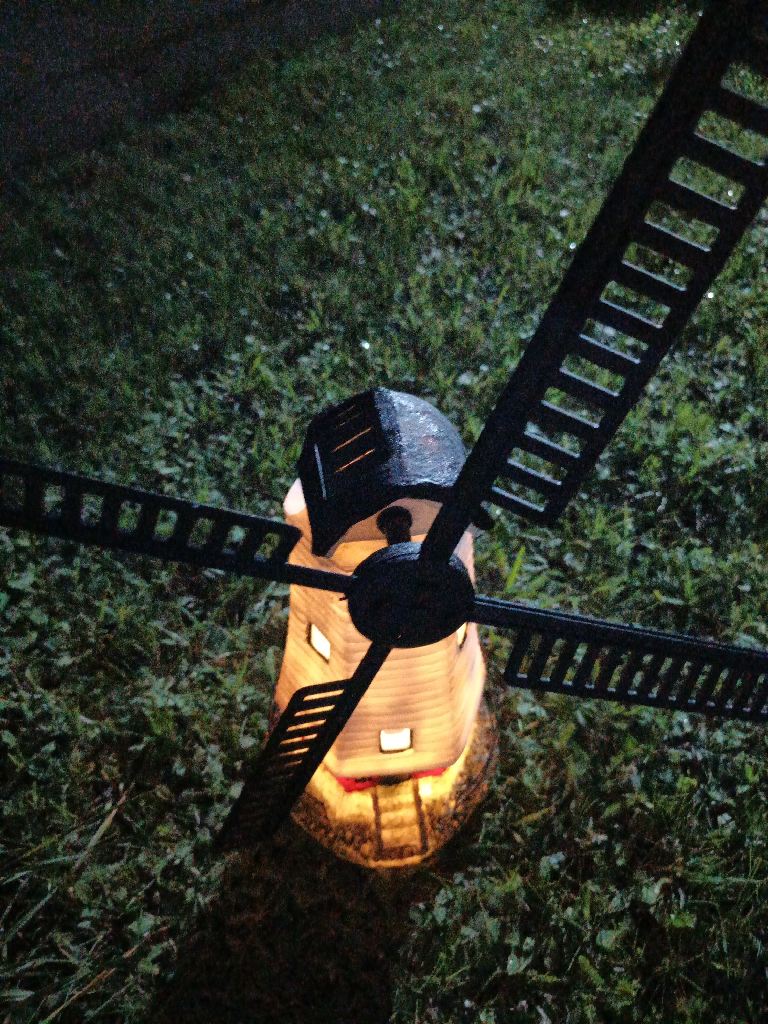 An illuminated windmill decoration sits on green grass in Лосева, Belarus. The windmill is made of a light-colored material and emits a warm, yellow light from its windows and base. It has a black, pointed roof and four black blades, each with a slatted design. The blades are positioned horizontally and diagonally relative to the windmill's body. The surrounding environment is dark, suggesting it is nighttime or dusk. The grass appears somewhat wet, with small reflective spots visible, possibly dew or raindrops. No people are present in the image.