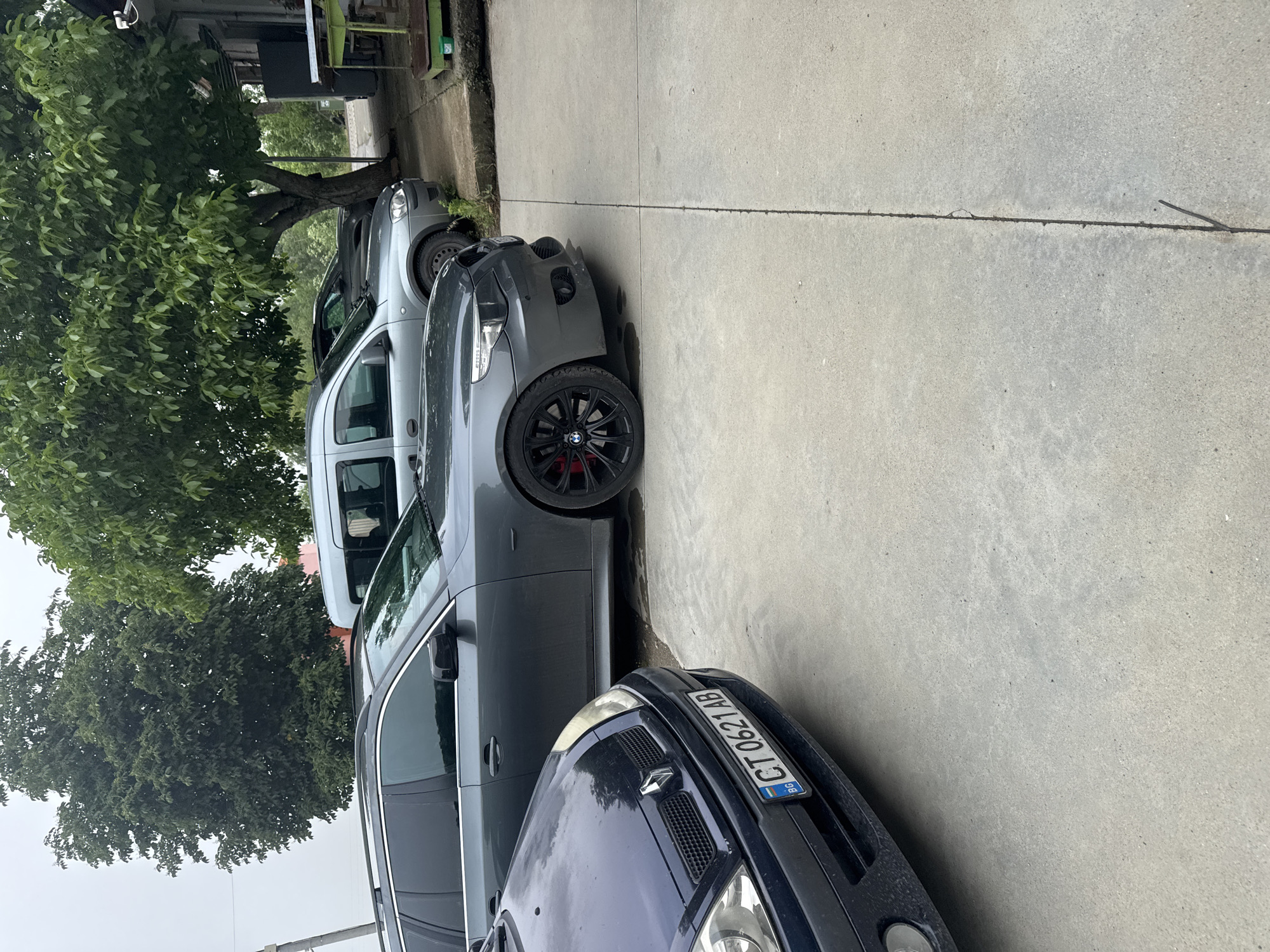 The image shows a row of four vehicles parked on a concrete surface, extending from the foreground into the midground. Lush green trees flank the parked cars on the left, with parts of a building visible in the background.
In the foreground, the front of a dark blue or black car, identified by its Renault badge, is visible. Its license plate clearly reads "CT 0621 AB" with a blue "BG" country code sticker, indicating Bulgarian registration. Directly behind this car is a dark grey vehicle, appearing to be a BMW, characterized by its black alloy wheels with a red accent on the brake calipers. Further down the line, a lighter grey or silver van-like vehicle is parked, followed by another light-colored car partially obscured by the foliage.
Large, leafy green trees dominate the left side of the frame, suggesting a verdant outdoor setting. A thick tree trunk is visible near the mid-section of the parked cars. In the upper left background, details of a building, including a roofline and a white fixture that resembles a security camera, can be seen.
The scene takes place during the daytime, under what appears to be an overcast sky, providing soft, diffused lighting. The concrete ground features visible seams and minor irregularities. No people are present, and the cars are stationary. The visible "BG" on the license plate aligns with the location context of Sarnevo, Bulgaria.