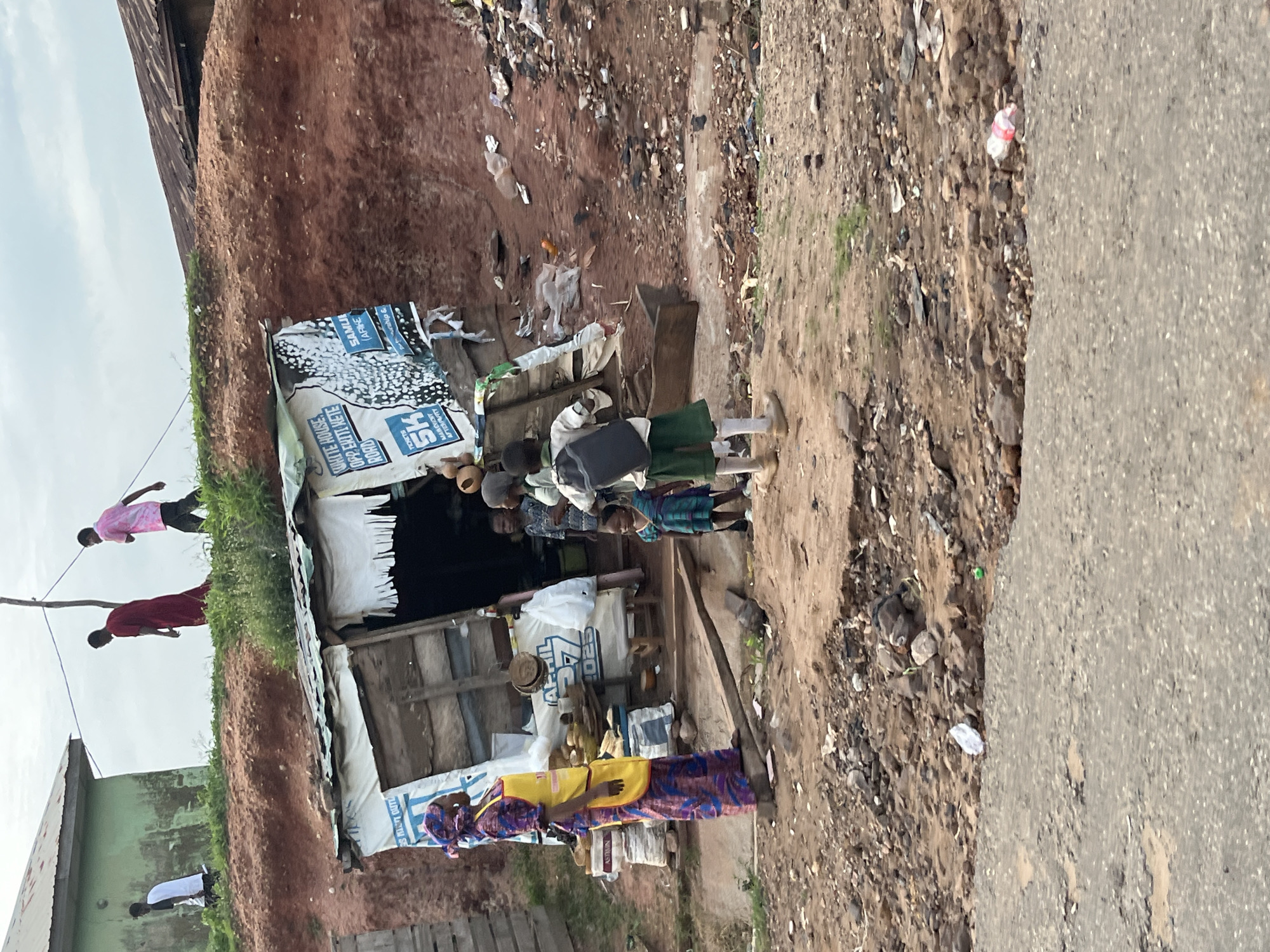The image depicts an outdoor scene in what appears to be a less developed area. In the foreground, a dirt road with some debris is visible, transitioning into a rough, rocky terrain. Behind the road, a small, makeshift stall or shop is set up against a reddish dirt embankment. The stall has a corrugated metal roof and appears to be constructed from salvaged materials. Signs are attached to the stall, one with text in English and another with what seems to be a graphical design.
Several individuals are present around the stall. A woman in a colorful patterned dress and a yellow apron stands to the left of the stall, facing away from the camera. To her right, three children are gathered near the stall entrance. One child, wearing a green and blue outfit, stands closer to the foreground. Another child, wearing a white shirt and green shorts with a backpack, is positioned further back, looking towards the stall. A third, smaller child, also in a colorful outfit, is partially visible behind the child in green and blue.
In the background, on top of the embankment, three figures are walking away from the camera. The figure on the left wears a white shirt and dark shorts. The middle figure is dressed in a red robe, and the figure on the right wears a pink and white tie-dye shirt and dark pants. A green building with a dark roof is situated on the far left of the embankment, and more dilapidated structures are visible in the distance to the right. The sky is overcast. The scene suggests a commercial or informal market setting in a rural or semi-urban environment, with elements of poverty and informal housing visible.