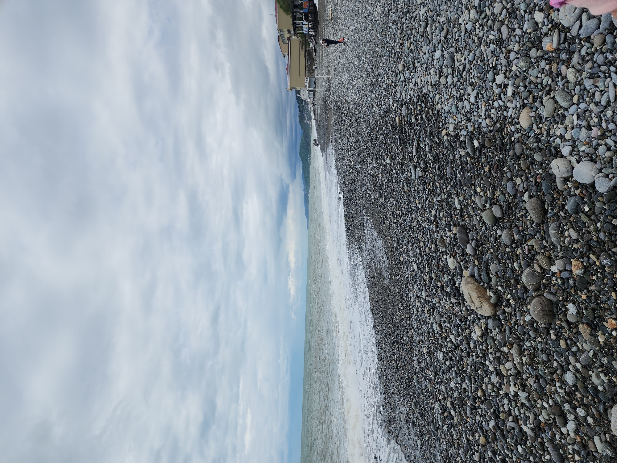 The image shows a pebble beach in Sochi, Russia. The sky is cloudy. The sea is calm with small waves lapping the shore. Several buildings are visible in the background along the shoreline. A person is standing near the buildings on the beach.