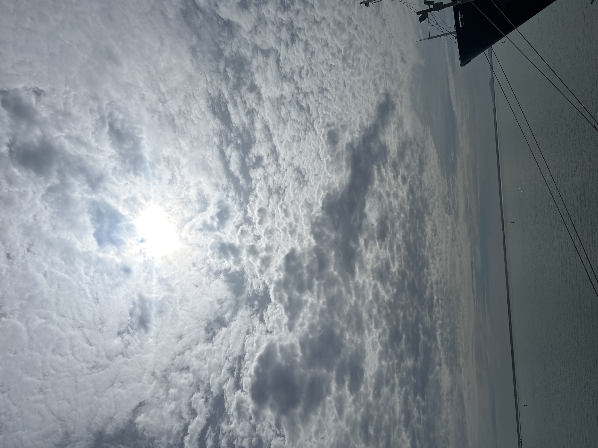 The image captures a coastal scene in Uribia, Colombia, under an overcast sky during daytime. The upper two-thirds of the frame are filled with a dense formation of altocumulus clouds, through which the sun is visible as a bright, diffused light source in the upper center, creating a distinct glare. The cloud cover exhibits varying densities and textures, with darker, more pronounced cloud masses contrasting with lighter areas around the sun.
The lower third of the image features a calm body of water, likely a bay or sea, reflecting the grey tones of the sky. Two small, red-topped navigational buoys are present on the water's surface: one in the lower-left quadrant and another in the lower-right, closer to the distant horizon. A low-lying, linear landmass or coastline stretches across the horizon, separating the water from the sky.
In the lower-right corner, the dark-colored bow of a large, stationary vessel is prominently visible, extending vertically into the frame. Multiple thick mooring lines originate from the vessel and descend diagonally towards the bottom of the image. A portion of the ship's superstructure, including a mast or antenna with attached fixtures, is also visible above the bow. No individuals are present in the depicted scene.