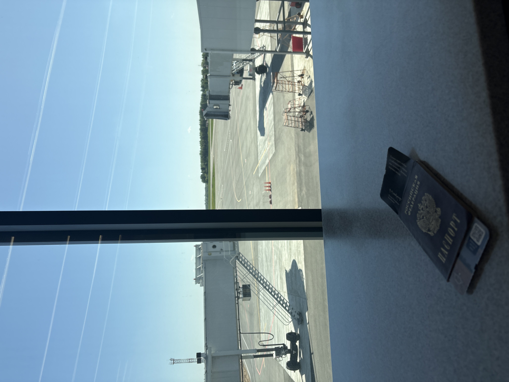 A view from a window at what appears to be an airport. Outside the window, there's an airport tarmac visible, with service vehicles and jet bridges. Above the tarmac is a blue sky streaked with contrails. In the foreground on the right, a Russian passport is placed on a flat surface, likely a table.
The airport is likely located in Новое Мартьяново, Russia.