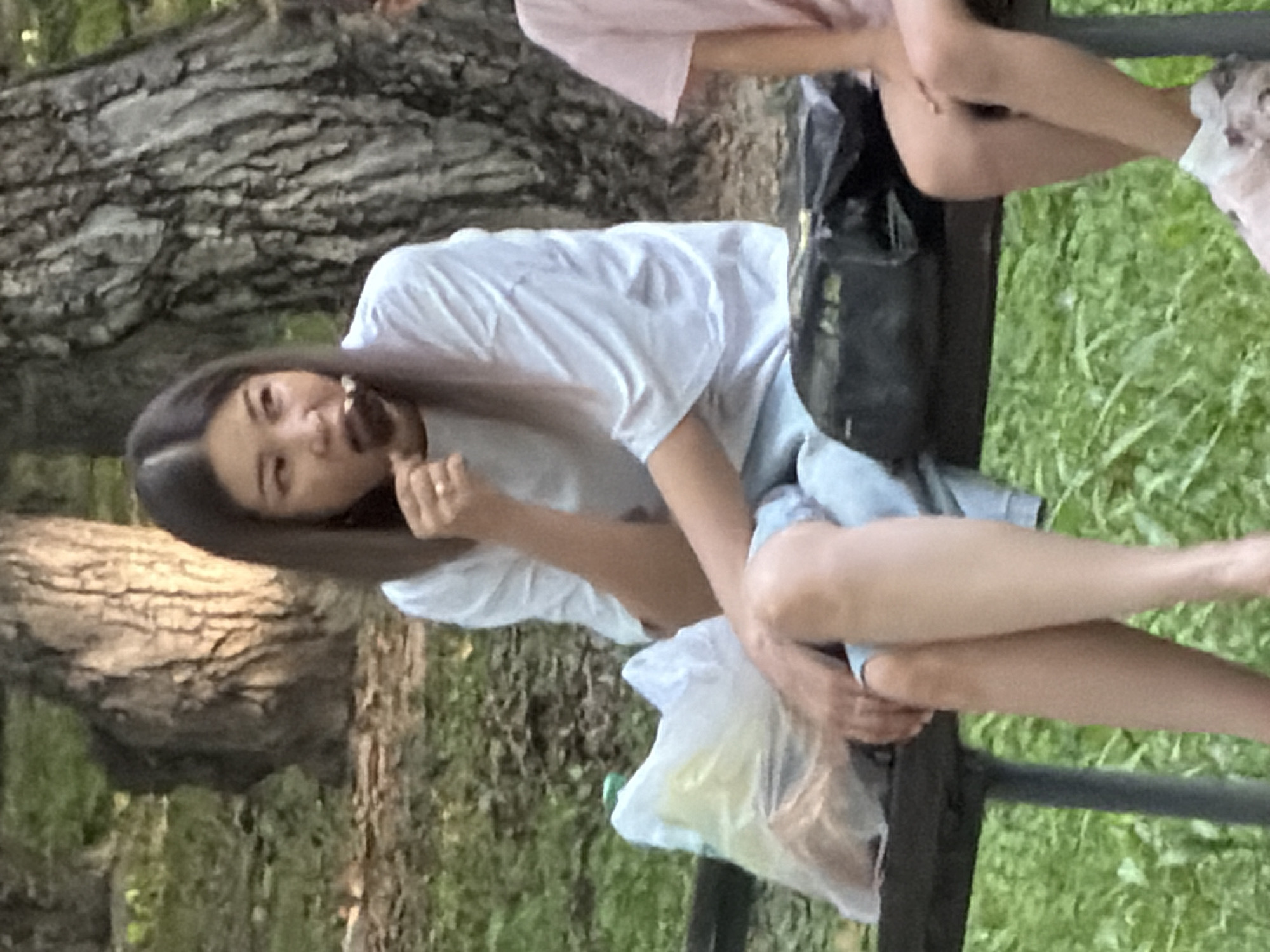 A woman with dark hair is sitting on a park bench and eating an ice cream. She is wearing a white t-shirt and blue shorts. There is a plastic bag with food next to her on the bench. Another person, likely a child, wearing a pink t-shirt and pink shoes, is sitting to her right. The background shows trees and grass, indicating an outdoor park setting.