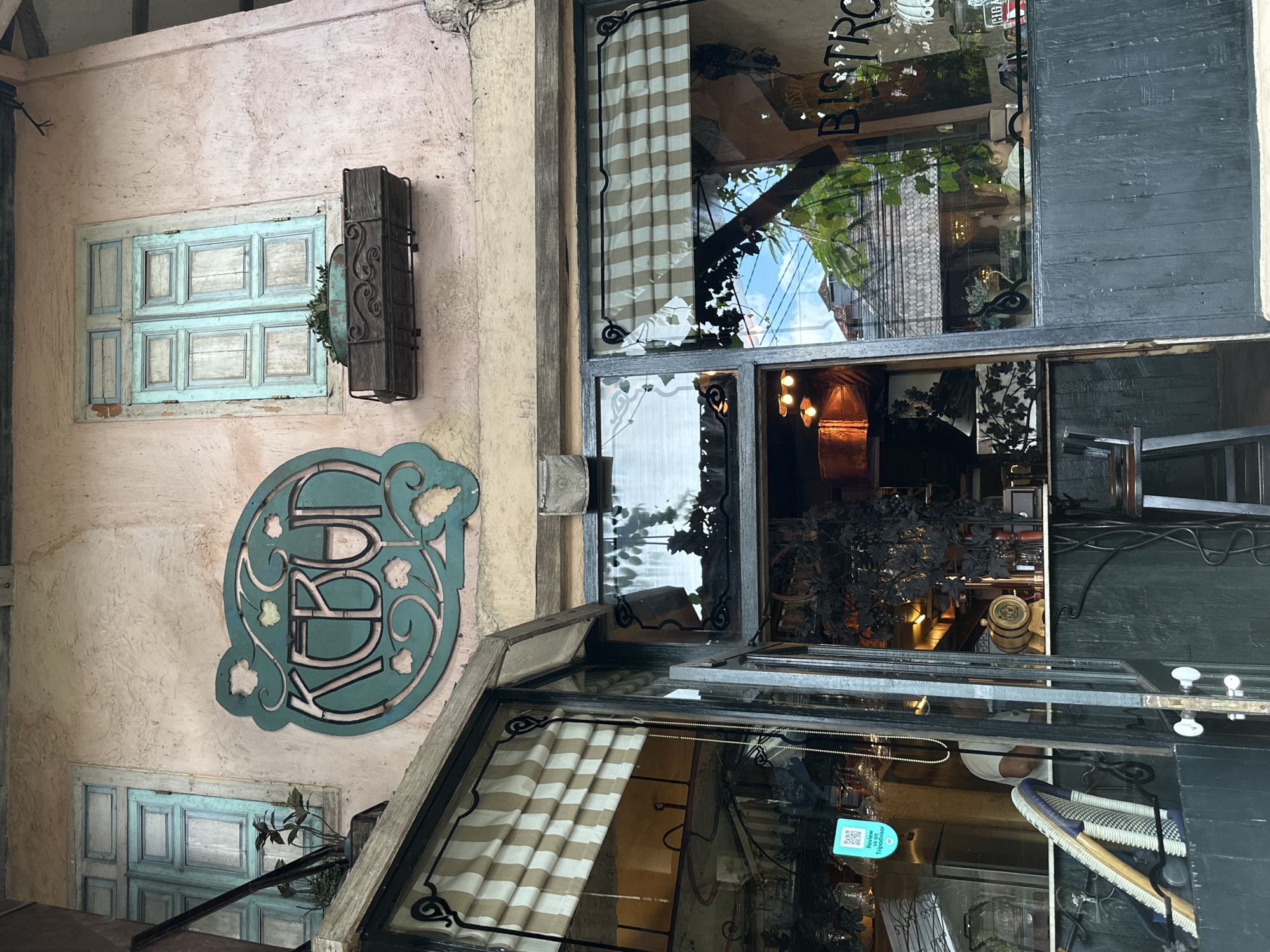 The image depicts the exterior of a restaurant or bistro named "KEBUN" in Ubud, Indonesia. The sign, in a stylized green font with vine motifs, is mounted on a pale pink stucco wall. To the left of the sign is a light blue wooden window with two panes, and a dark wooden planter box with greenery below it. The entrance to the establishment is visible, with a dark wooden facade and striped awnings. Through the glass doors and windows, the interior of the bistro can be seen, with dimly lit areas, tables, chairs, and decor. In the reflection on the window, buildings and trees are visible, suggesting an outdoor setting. A QR code is displayed on a sticker on the glass, likely for reviews or menus.