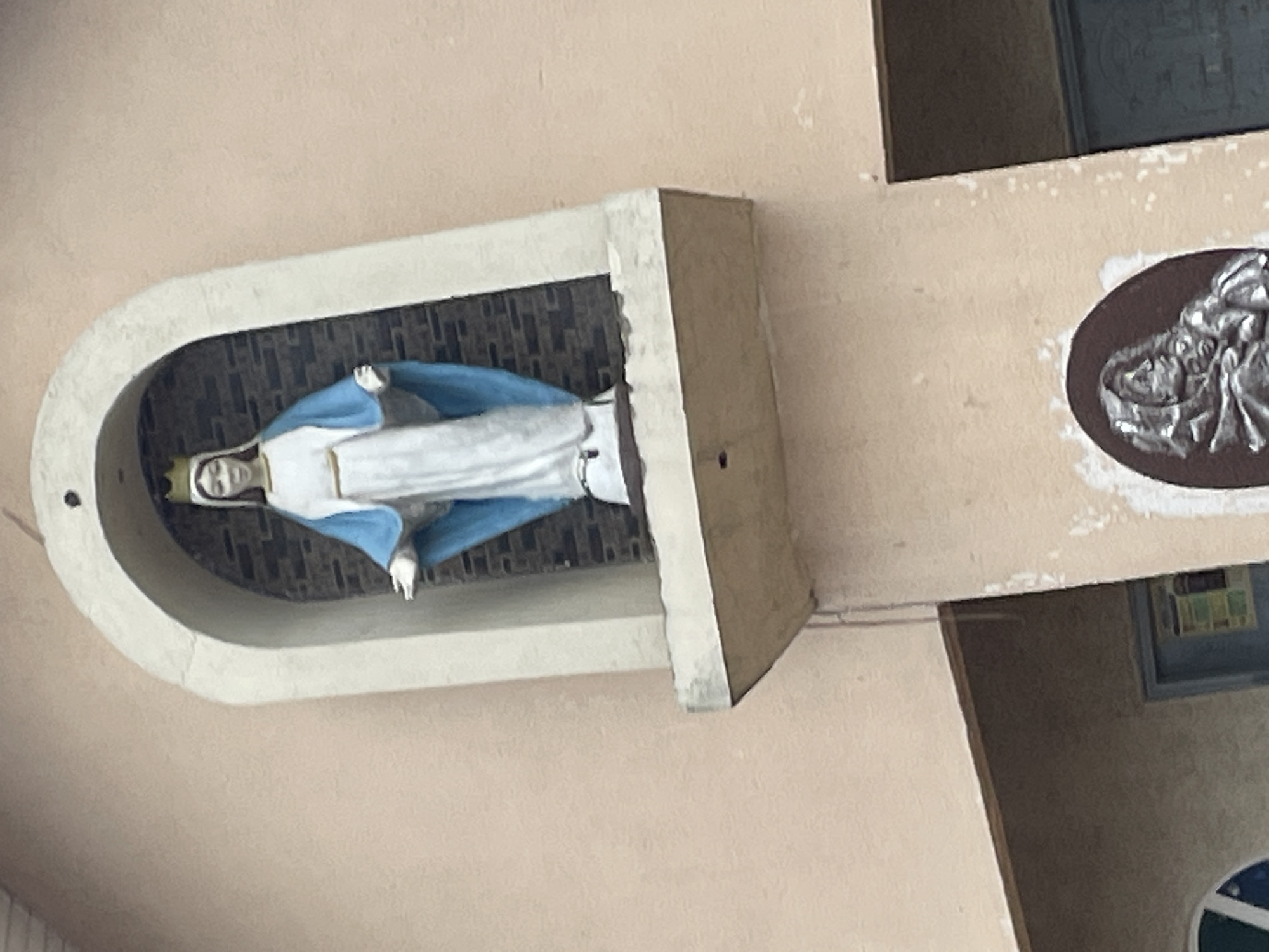 A statue of the Virgin Mary, adorned with a golden crown and a blue mantle over a white robe, stands within an arched niche on the exterior of a building. The niche is framed by a textured, light-colored material, and the background within the niche appears to be brickwork. Below the niche, a concrete ledge extends outwards, showing some signs of wear and tear. Further down on the building's facade, an oval-shaped relief carving is visible, depicting a robed figure with a hooded head. The building's exterior wall is painted a pale peach color with some areas of peeling paint and staining. To the left of the niche, part of a window with a blue pane is visible. To the right of the relief carving, a section of a blue doorway can be seen. This scene is located in Jos, Nigeria, specifically on the facade of the Catholic Church of the Sacred Heart.