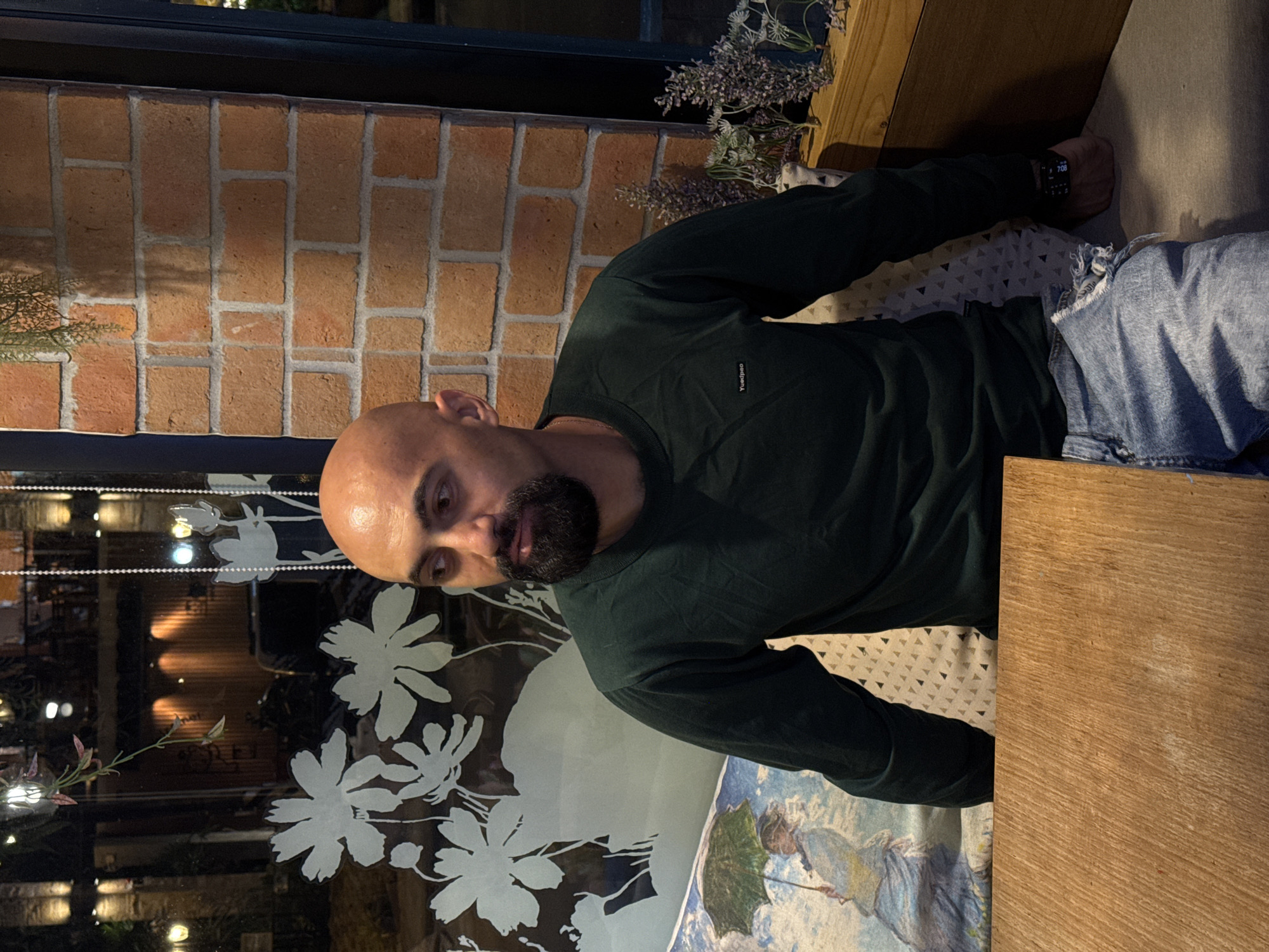 A bald man with a beard is seated at a wooden table in what appears to be a cafe or restaurant. He is wearing a dark green long-sleeved shirt and ripped blue jeans. The background includes a brick wall, a window with floral decals, and some potted plants. The scene is dimly lit, suggesting it might be evening or nighttime. The overall atmosphere is casual and relaxed. This scene is located in the city of Ko Samui District, Thailand.