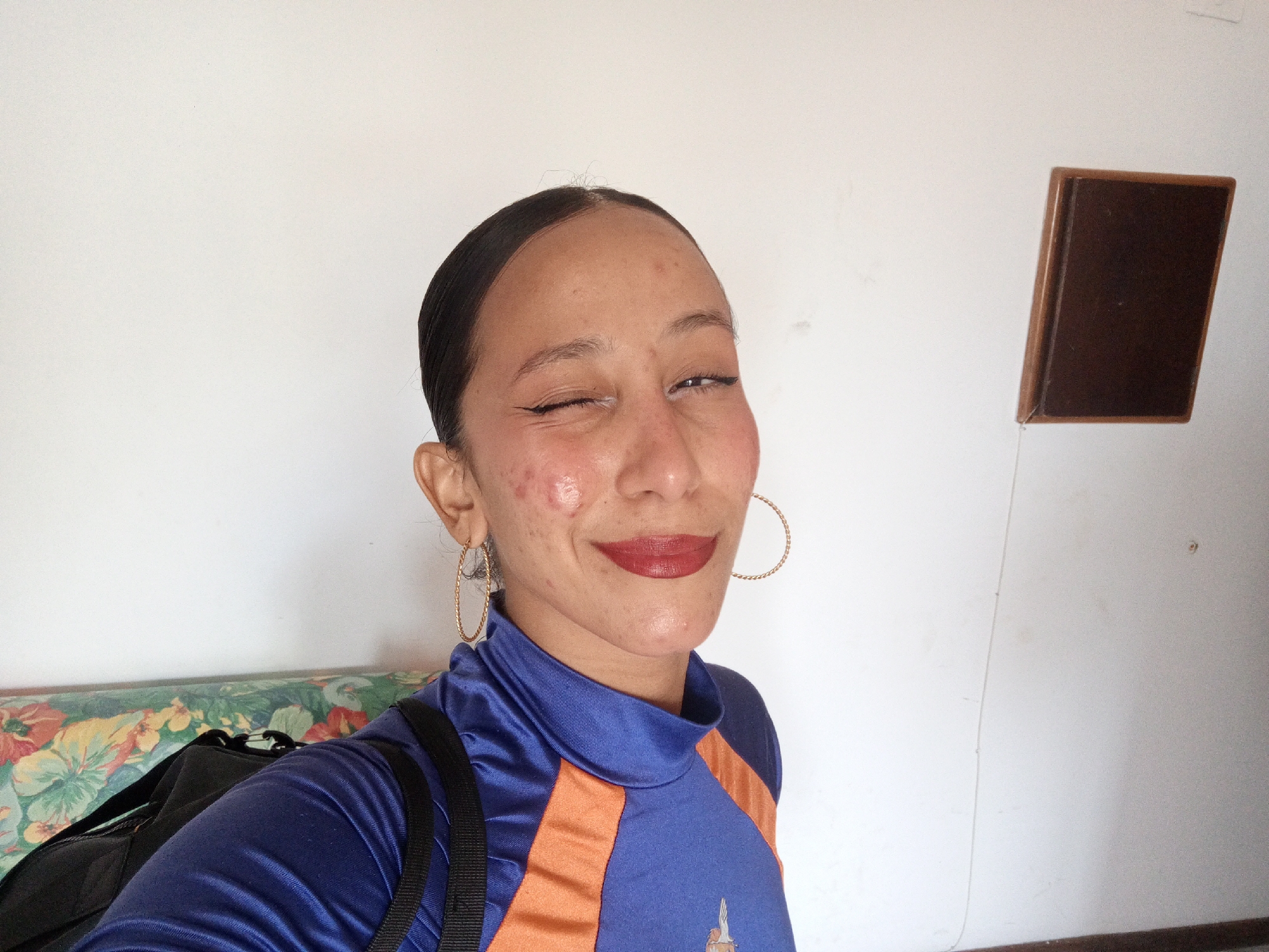 A medium-shot, selfie-style photograph shows a person with dark hair pulled back, wearing gold hoop earrings and a blue and orange athletic top. They are winking at the camera, with dark lipstick on. A black backpack with straps is visible over their left shoulder.
The background consists of a white wall, with a floral print couch to the left and a brown, rectangular object mounted on the wall to the right. The photograph appears to be taken indoors, possibly in a home.
There is no information available in this image to identify the city as Porlamar, Venezuela.