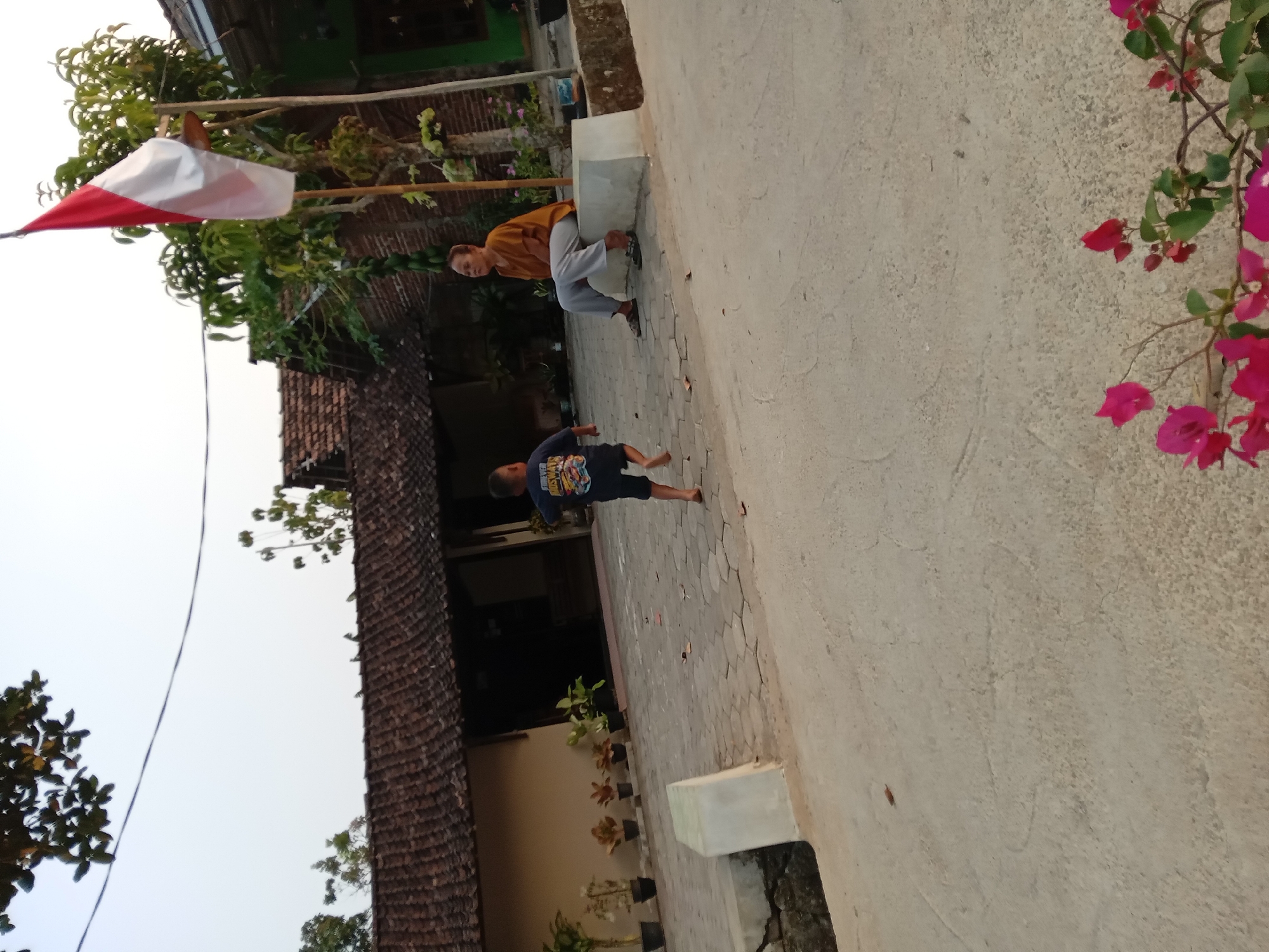The image depicts a scene in Kedunggudel, Indonesia, likely in a rural or semi-rural residential area. A young boy in a dark t-shirt and shorts is running across a paved courtyard. To the right, a woman wearing a mustard-colored shirt and grey pants is sitting on a low, concrete bench. Behind them, a traditional Indonesian flag, red and white, flies from a bamboo pole. The background features a building with a tiled roof, characteristic of Indonesian architecture, and several potted plants. The ground is paved with interlocking concrete blocks. In the foreground, a branch with pink bougainvillea flowers is visible. The overall lighting suggests it is either early morning or late afternoon.
