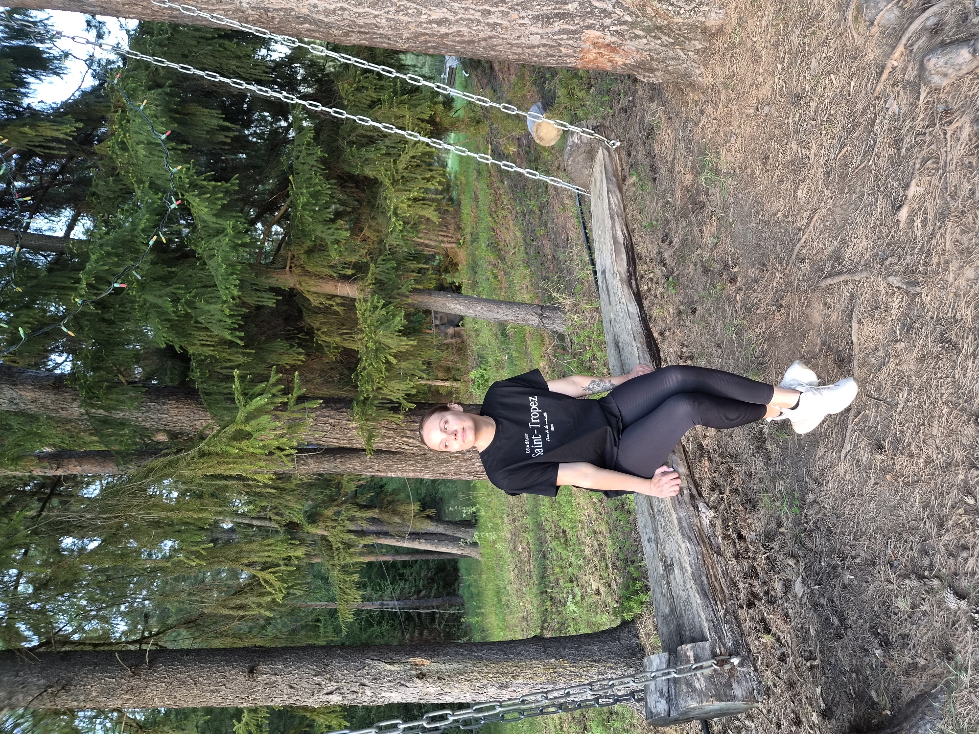 A full-length shot shows a woman sitting on a wooden bench swing suspended by chains between two trees. She is wearing a black t-shirt with "Saint-Tropez" printed on it, black leggings, and white sneakers. The woman is looking directly at the camera. The swing is located in a wooded area with tall evergreen trees and a ground covered in dry pine needles and grass. The location is Прикамский, Russia.
