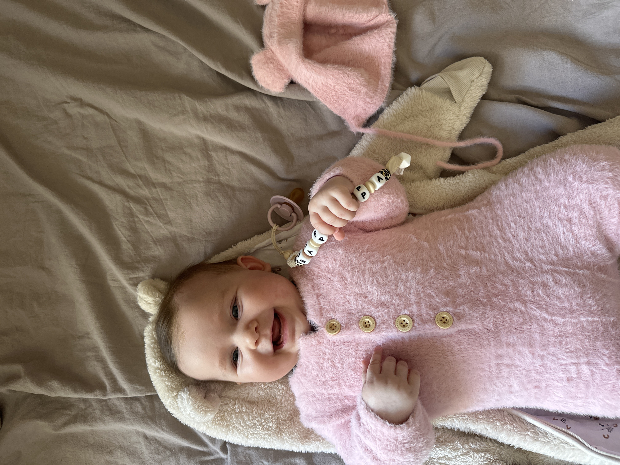 A baby lies on a gray bed sheet, wearing a pink furry jumpsuit with wooden buttons and a beige plush headrest with small ears. The baby is holding a pacifier clip with alphabet beads in its left hand and smiling. A pink furry hat and a pacifier are near the baby. The environment appears to be indoors. It is impossible to determine the location in Chaikovsky, Russia from the image.