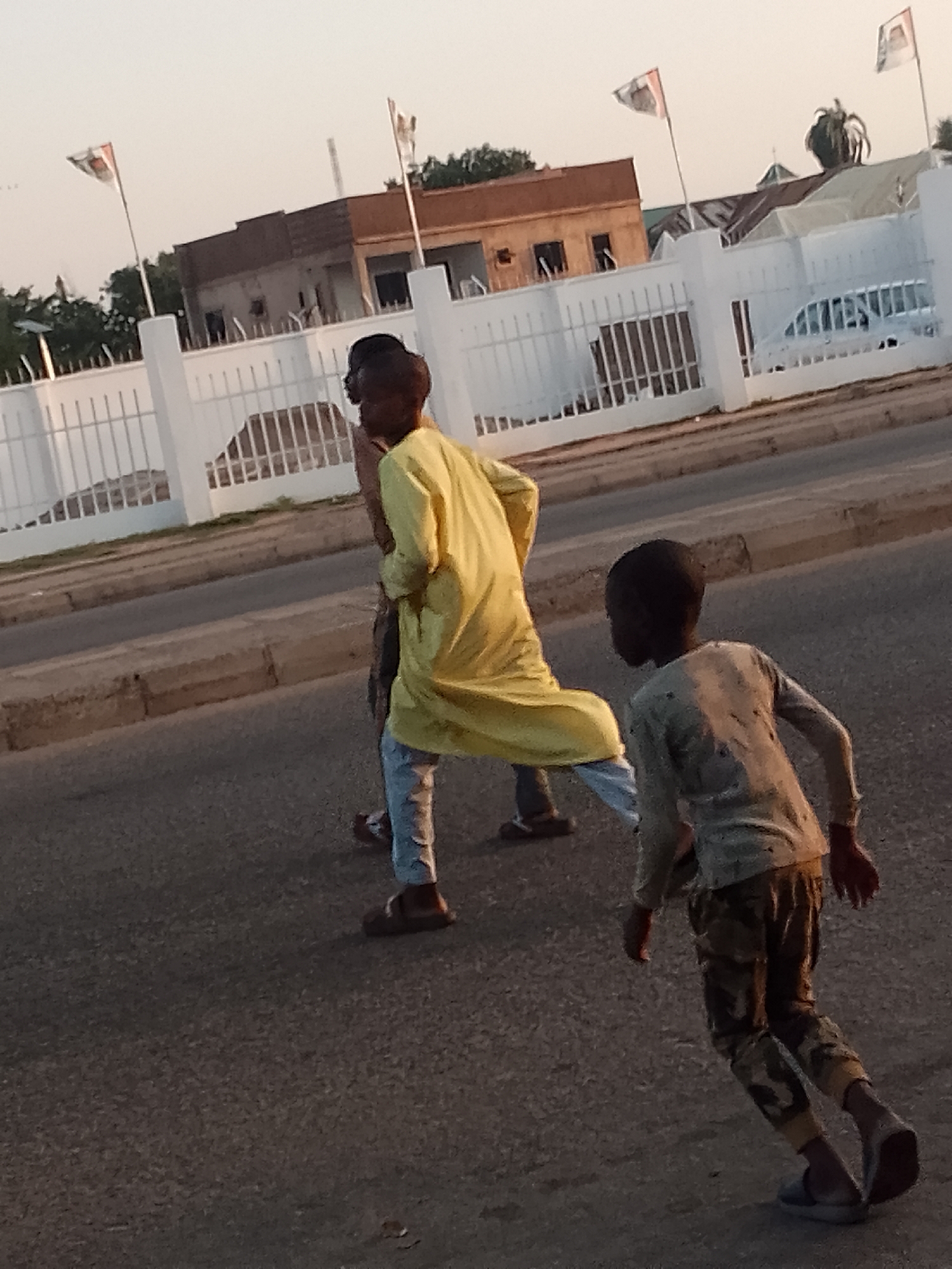 The scene depicts two individuals on an asphalt road in Pantame, Nigeria. The individual in the foreground, a child, is moving rightward in a running posture, wearing a light-colored, long-sleeved shirt with dark stains and camouflage-patterned trousers with light-colored slip-on shoes. The second individual, an adolescent or young adult, is located behind and to the left of the first, also moving rightward. This individual wears a long yellow tunic over light-colored trousers and dark sandals. In the background, a white perimeter fence with vertical metal bars separates the road from a property containing several buildings and vegetation. Two white rectangular flags, each featuring a distinct design, are visible on poles above the fence line. A light-colored vehicle is partially visible parked behind the fence on the right. The sky is clear, and the lighting suggests daylight conditions.