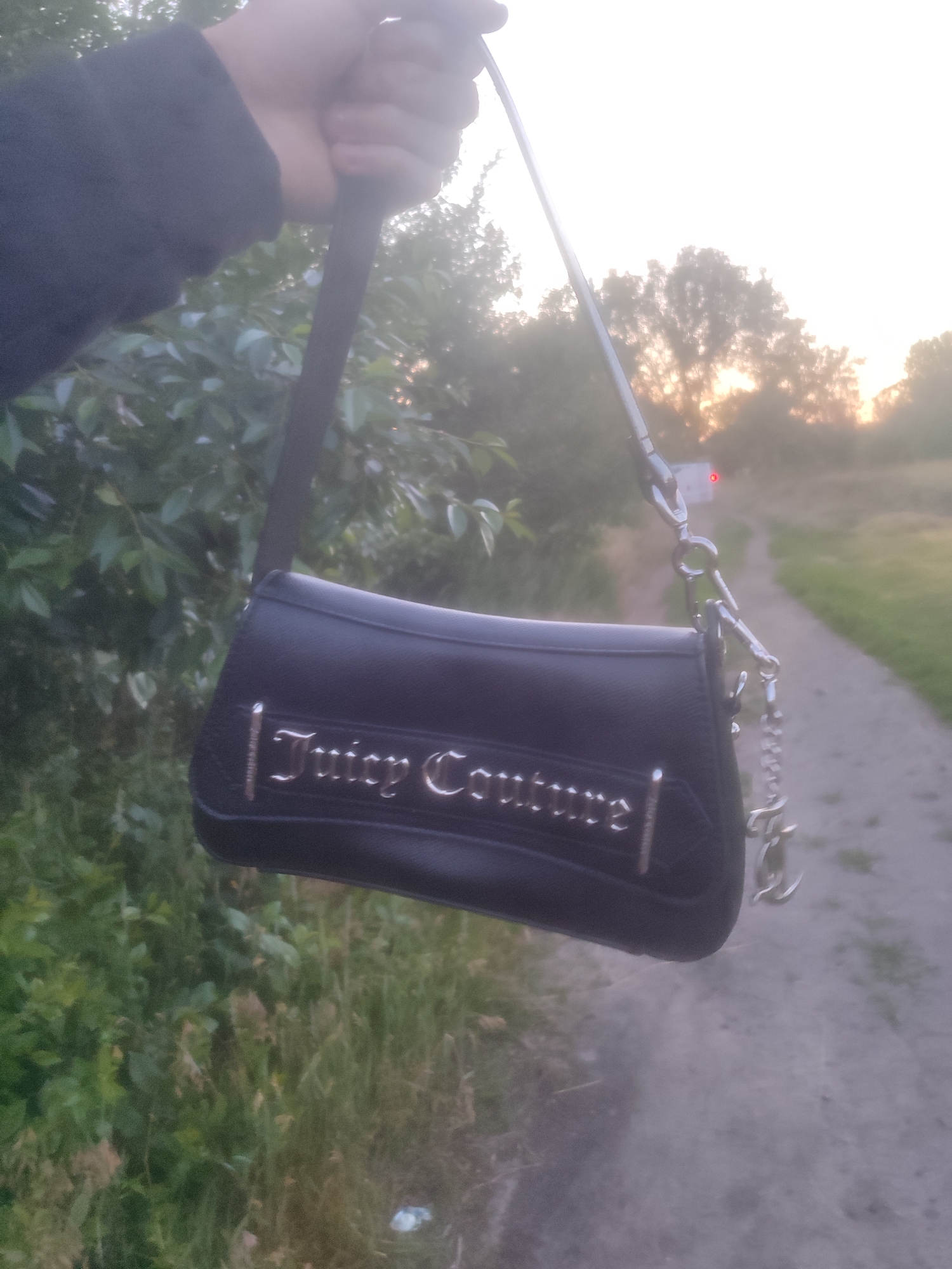 A person is holding a black Juicy Couture purse with a silver chain strap. The purse has the brand name "Juicy Couture" written in silver on the front. The person is wearing a dark sleeve. The purse is being held in front of a background of trees and a dirt path. The sun appears to be setting or rising, casting a warm glow on the scene. The location is Unknown, Unknown.