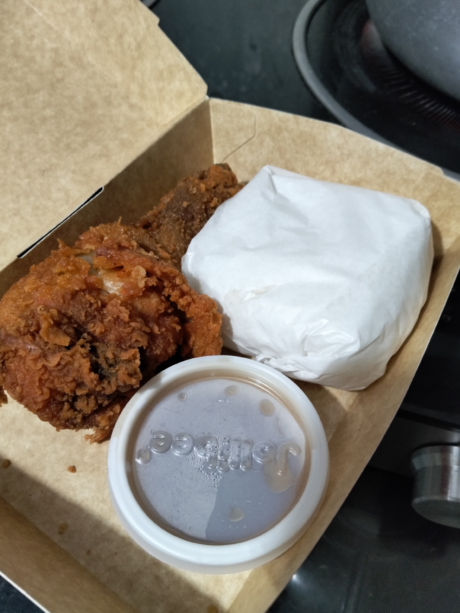 This is a high-angle, close-up, eye-level shot of a Jollibee meal box in Davao City, Philippines. The box is made of light brown cardboard. Inside the box, there is a piece of crispy fried chicken, a white paper-wrapped item, and a small white plastic container with a lid. The lid of the container has the embossed Jollibee logo. The fried chicken appears to be a leg or thigh piece, with a thick, golden-brown crispy coating. The white paper-wrapped item is likely a side dish or another piece of food. The container is filled with a dark liquid, possibly gravy or sauce, and some small bubbles are visible on the surface. The background is dark and out of focus, showing a portion of a black stovetop or counter. The lighting is artificial and comes from above, creating some shadows within the box.