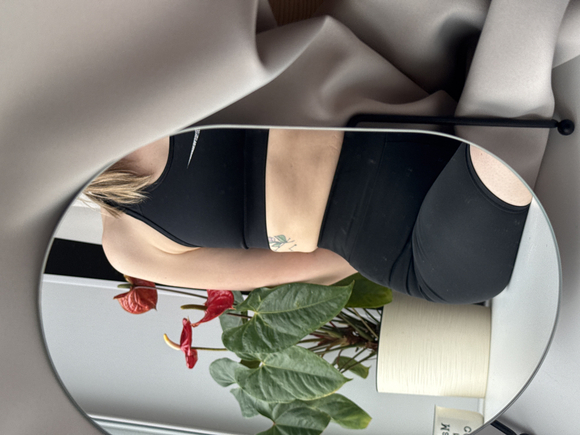 A mirror selfie captures a person's torso and an indoor plant in a minimalist indoor setting. The central subject is the reflection in a round mirror, showing a person, likely female, from the chest to the upper thigh. They wear a black sports bra with a subtle white logo on the left strap and black athletic shorts or leggings. Blonde or light brown hair is visible near their shoulder. A small green tattoo, resembling a leafy design, adorns their left side/torso.
Within the mirror's reflection, an indoor plant with vibrant red anthurium flowers and large green leaves is prominently featured. It rests in a light-colored, textured cylindrical pot. The mirror itself is supported by a thin, black metal stand with a spherical end, visible behind the person in the upper right area. Outside the mirror's frame, behind the person, a soft light gray or beige draped fabric creates a textured backdrop. The background reflected in the mirror is a plain light gray or white wall. In the bottom right corner of the mirror's reflection, partially visible text reading "SM" and "Ms" can be seen on a small, dark object. The overall scene suggests a stylish interior, where the person is posing for a photo, showcasing their athletic wear and tattoo.