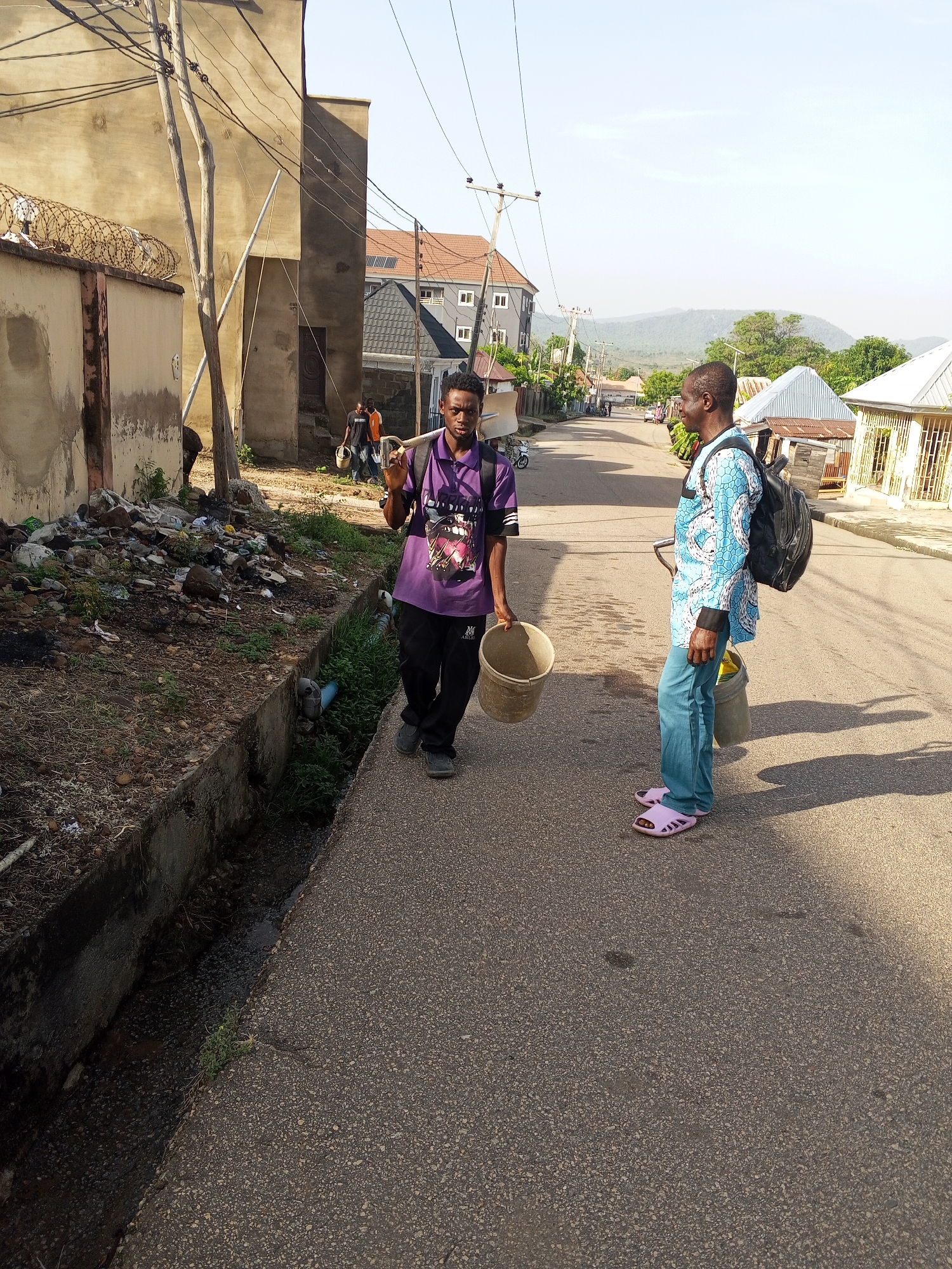 The image depicts a street scene in Lokoja, Nigeria. Two men are walking on a paved road. The man on the left wears a purple shirt and black pants, holding an empty bucket. The man on the right wears a blue patterned shirt and blue jeans, with pink sandals. He also carries a bucket and wears a backpack. The background includes buildings of varying designs, power lines, and a distant hillside. To the left, there is a drainage ditch with accumulated trash next to a concrete wall topped with barbed wire.
