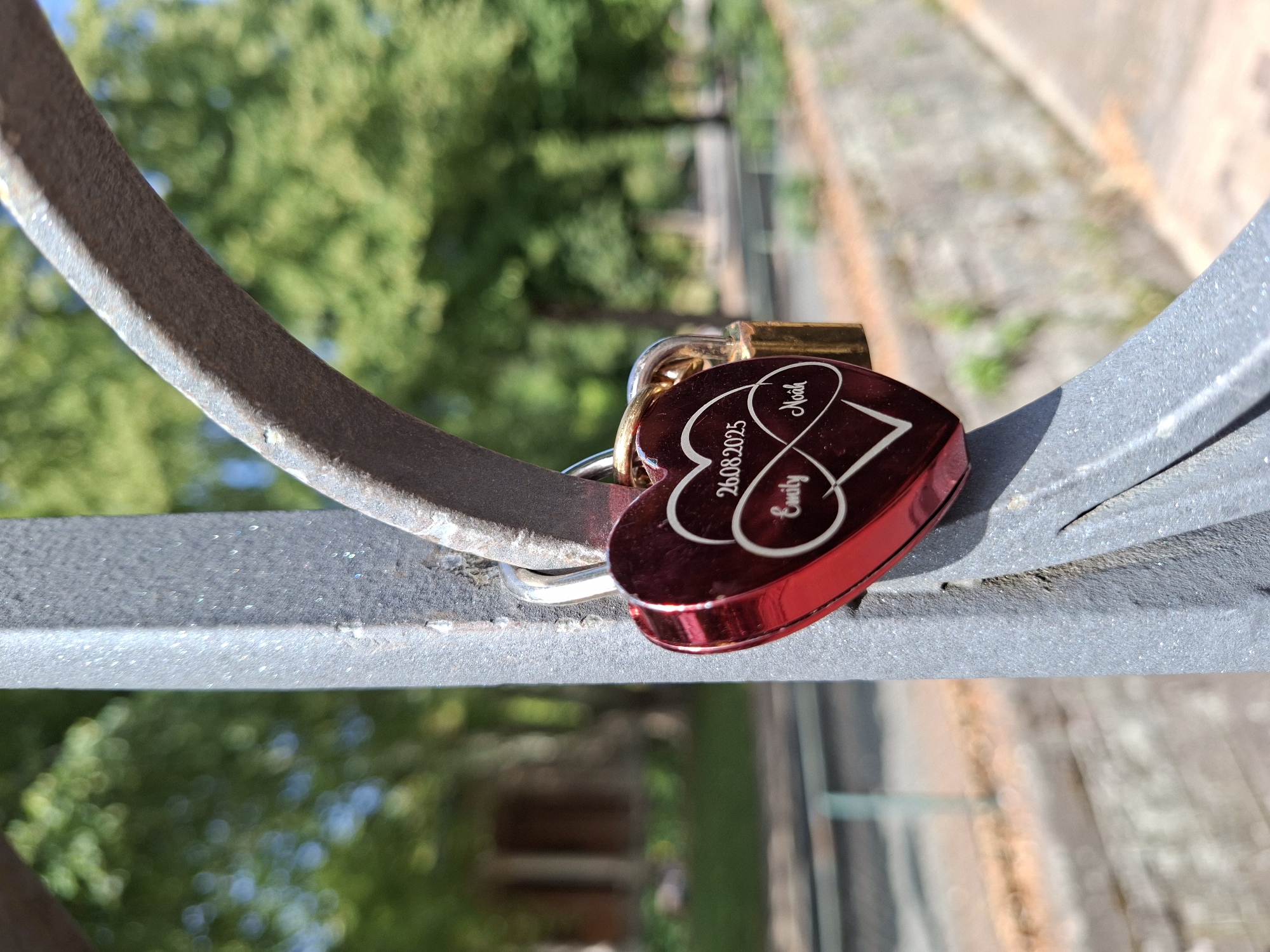 The image shows a close-up of a red heart-shaped padlock attached to a metal railing, likely on a bridge, in Baden-Baden, Germany. The padlock has an infinity symbol engraved on it, along with the names "Emily" and "Noah" and the date "26.08.2025". A smaller gold padlock is attached to the same railing, above the heart-shaped lock. The background is blurred, showing trees with green foliage and a hint of blue sky, suggesting an outdoor, park-like setting within the city. The railing appears to be made of weathered metal.