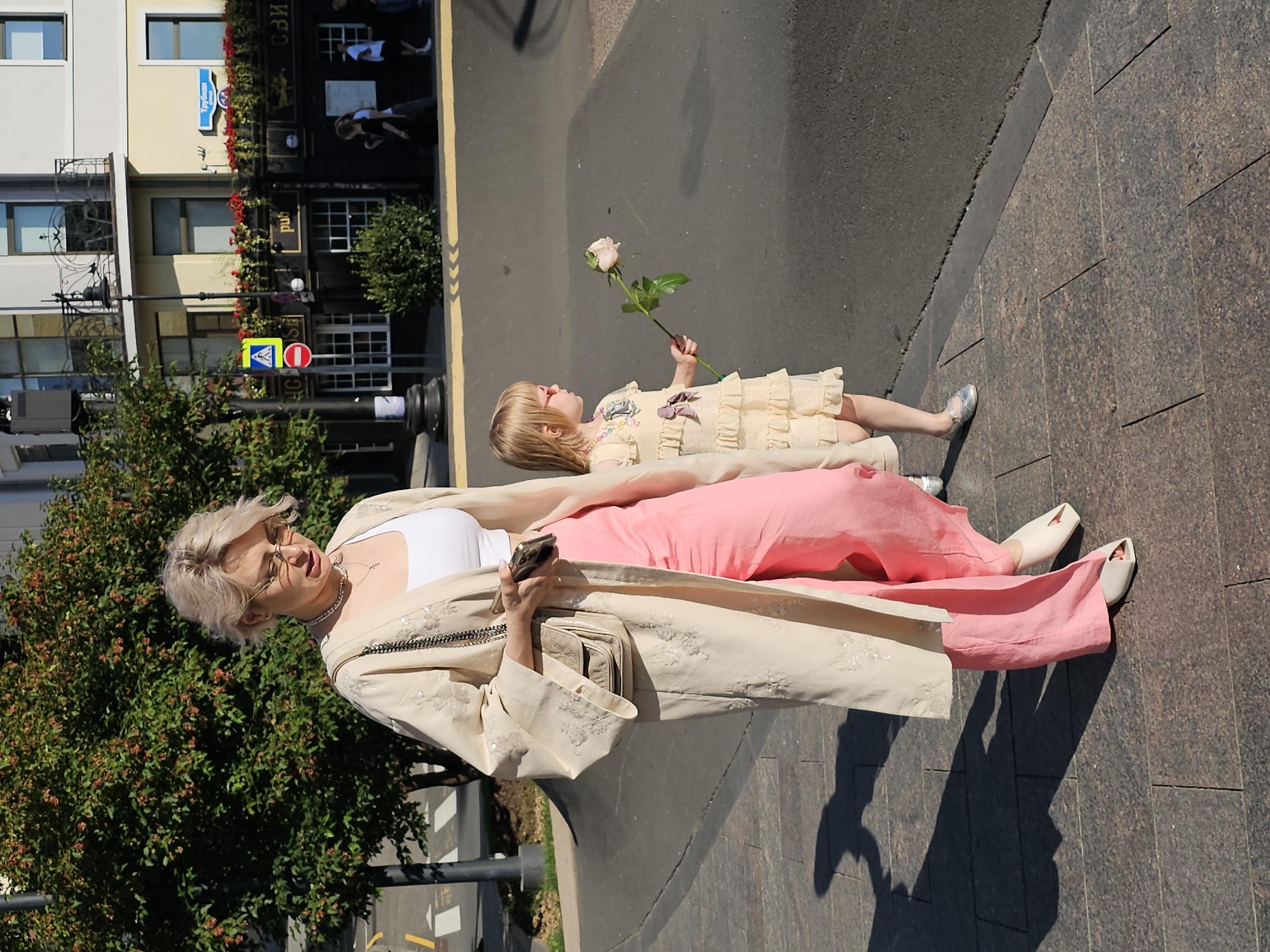 A woman with blonde hair and glasses, wearing a cream-colored kimono-style jacket over a white top and pink pants, walks on a sidewalk. She is holding a smartphone in her right hand and a light-colored purse is slung over her left shoulder. Beside her, a young girl with blonde hair, wearing a light yellow ruffled dress and pink sunglasses, walks while holding a single light pink rose. They are walking on a cobblestone sidewalk in front of a building with a pub sign that reads "pub" and "сви" (likely the beginning of a Russian word). The building is located on a street in Moscow, Russia, as indicated by the street sign with Cyrillic text. Trees and urban elements like a traffic light and pedestrian crossing are visible in the background. The scene is illuminated by bright sunlight, casting shadows on the ground.