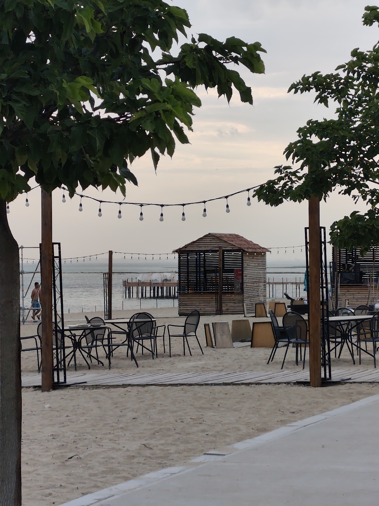 This image captures a serene beachside scene in Aktau, Kazakhstan. The foreground features an outdoor seating area with tables and chairs set up on a sandy beach. Wooden walkways provide a path through the sand. Beyond the seating area, several small wooden huts, possibly for vendors or changing rooms, are visible. A string of festoon lights hangs between wooden posts, suggesting an evening setting.
In the middle ground, the calm sea stretches out towards a distant pier with structures on it. A lone man, wearing blue shorts, is walking on the beach near some scaffolding. The background is dominated by a pale, overcast sky, indicating a peaceful, possibly cool day. The overall mood is one of tranquility and relaxation, perfect for a seaside resort. The location's context as Aktau, Kazakhstan, places this scene along the Caspian Sea.