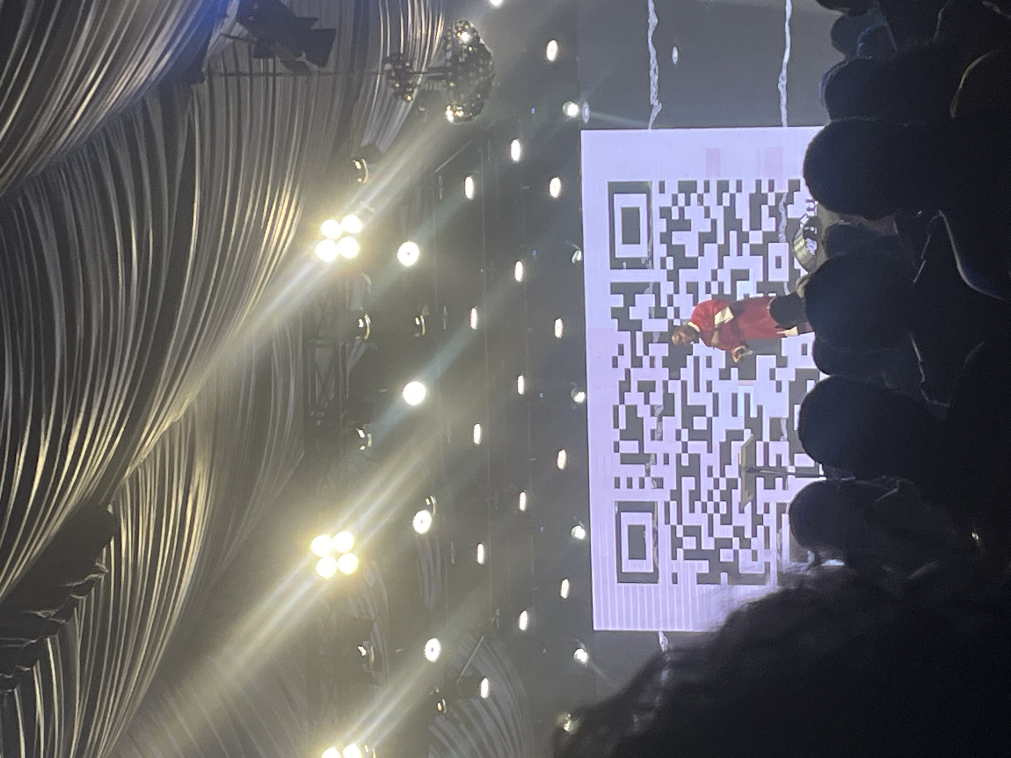 The image shows a stage setup, possibly an event or performance, with ornate gray drapery visible on the left side. Numerous stage lights are illuminating the scene. A large screen displays a QR code. In the middle of the screen, a person in a red shirt and black pants is visible. On the right side, part of the audience is visible in silhouette. The location is possibly Galadimawa Village, Nigeria.