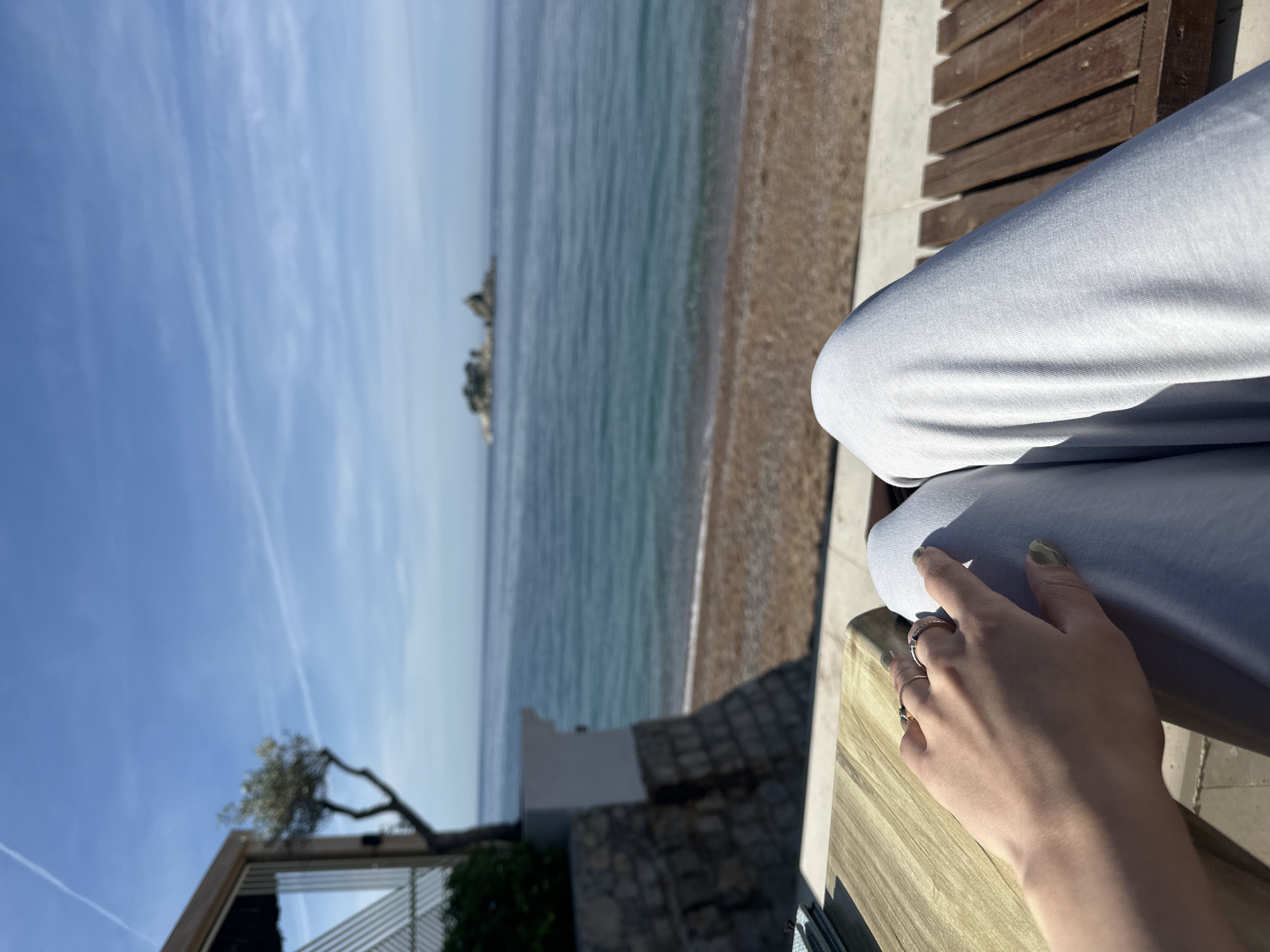 The photograph depicts a view from a seated position at a seaside location, likely Petrovac Centar, Montenegro, given the location's specific mention.
The frame is dominated by a pair of legs clad in light gray casual wear, resting on a wooden structure. A hand, adorned with rings and showing light green nail polish, is positioned on one of the legs.
In the background, a beach merges into a body of water, potentially the Adriatic Sea, under a sky with some clouds. In the distance, a rocky islet is visible.
To the left, part of a building structure is visible, with a tree branch extending from it towards the sky. The scene appears to be taken on a sunny day, giving the impression of relaxation in a beach setting.