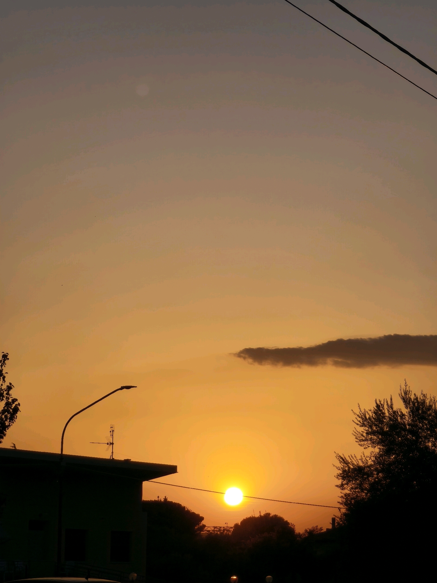 The image captures a sunset over Passignano sul Trasimeno, Italy. A bright, round sun is positioned low in the sky, casting a warm orange glow that dominates the scene. Silhouetted against the vibrant sky is a low-rise building with a prominent street lamp extending from its roof. Behind the building and to the right, a leafy tree is also silhouetted. A dark, elongated cloud drifts across the upper right portion of the sky, contrasting with the otherwise clear expanse. Power lines are visible against the gradient of the sky. The lower portion of the image is darker, with the silhouettes of trees and some indistinct shapes that may be vehicles. The overall impression is one of a tranquil evening in a suburban or semi-urban environment.