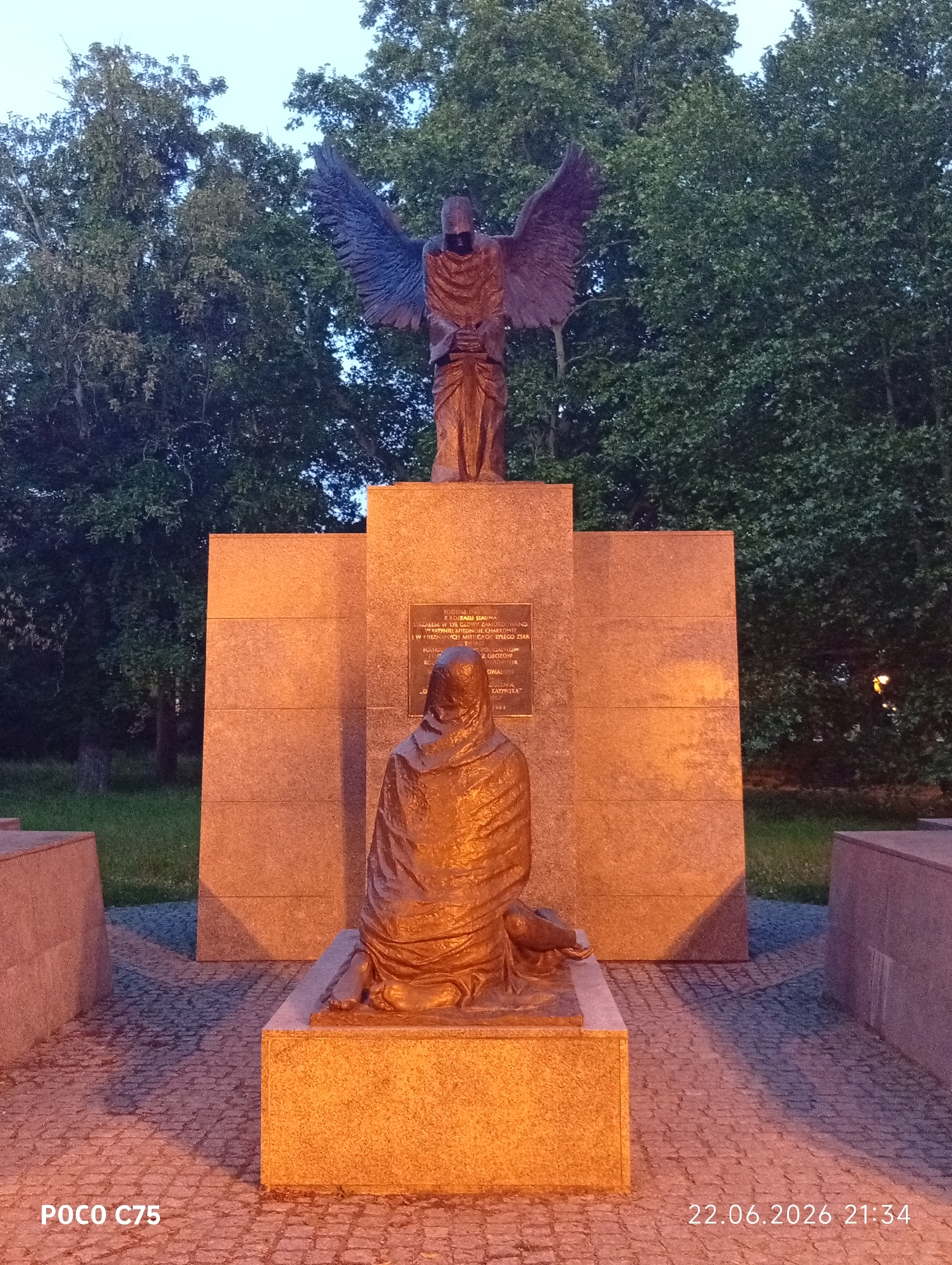 The image depicts a memorial statue in Wrocław, Poland, likely located in a park or public square. The central feature is a sculpture of an angelic figure with large wings, standing behind a seated figure draped in robes. The seated figure is positioned in front of a large, rectangular stone monument with inscribed text. The monument and the base of the statues are illuminated by artificial light, casting a warm glow. The background consists of lush green trees, suggesting an outdoor setting, possibly at dusk or dawn given the twilight sky visible at the top. The ground surrounding the monument is paved with cobblestones. The bottom of the image displays a watermark with "POCO C75" and the date "22.06.2026 21:34".