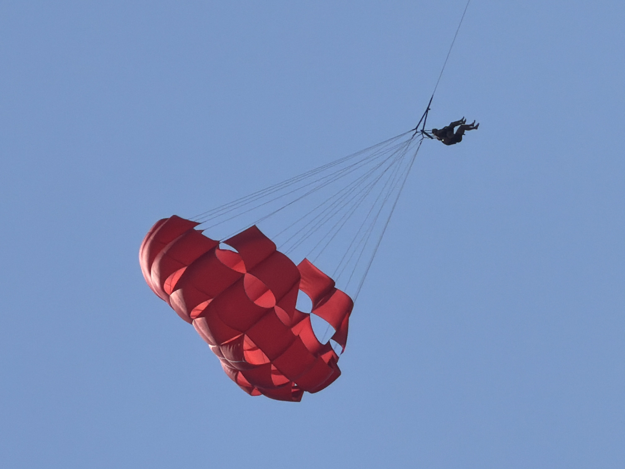 A vivid red ram-air parasail with two occupants is depicted against a clear, light blue sky, suggesting a daytime scene in Budva, Montenegro. The parasail, prominently positioned in the upper-left quadrant, is fully inflated and consists of multiple interconnected rectangular cells, giving it a characteristic winged shape. Its color is a uniform bright red, with subtle variations in shading indicating the contours of its cells. Numerous thin suspension lines extend from the parasail canopy, converging to a dark-colored tandem harness. Two individuals, appearing as silhouettes due to distance and angle, are seated side-by-side within this harness. They are dressed in dark attire, with legs visible and bent. A single, thicker black tow rope extends horizontally from the harness to the right side of the frame, indicating the activity of parasailing, where the individuals are being pulled by an unseen vessel. The background is entirely an unblemished, consistent shade of blue sky, devoid of clouds or other atmospheric features.