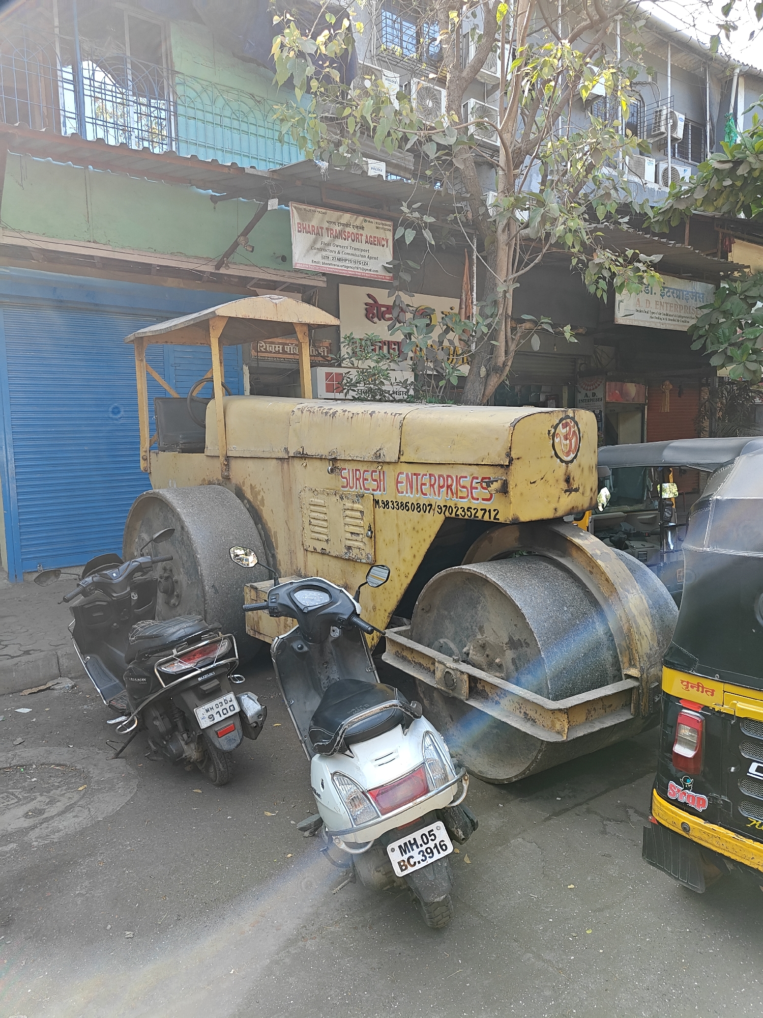 The image shows a yellow road roller parked on the side of a street in Mumbai, India. The road roller has the name "SURESI ENTERPRISES" painted on the side, along with a phone number. Two scooters and a yellow and black auto-rickshaw are parked next to the road roller. Buildings are visible in the background, with signs for various businesses, including "BHARAT TRANSPORT AGENCY" and "A. D. ENTERPRISES". There are trees lining the street, and the sky appears to be overcast.