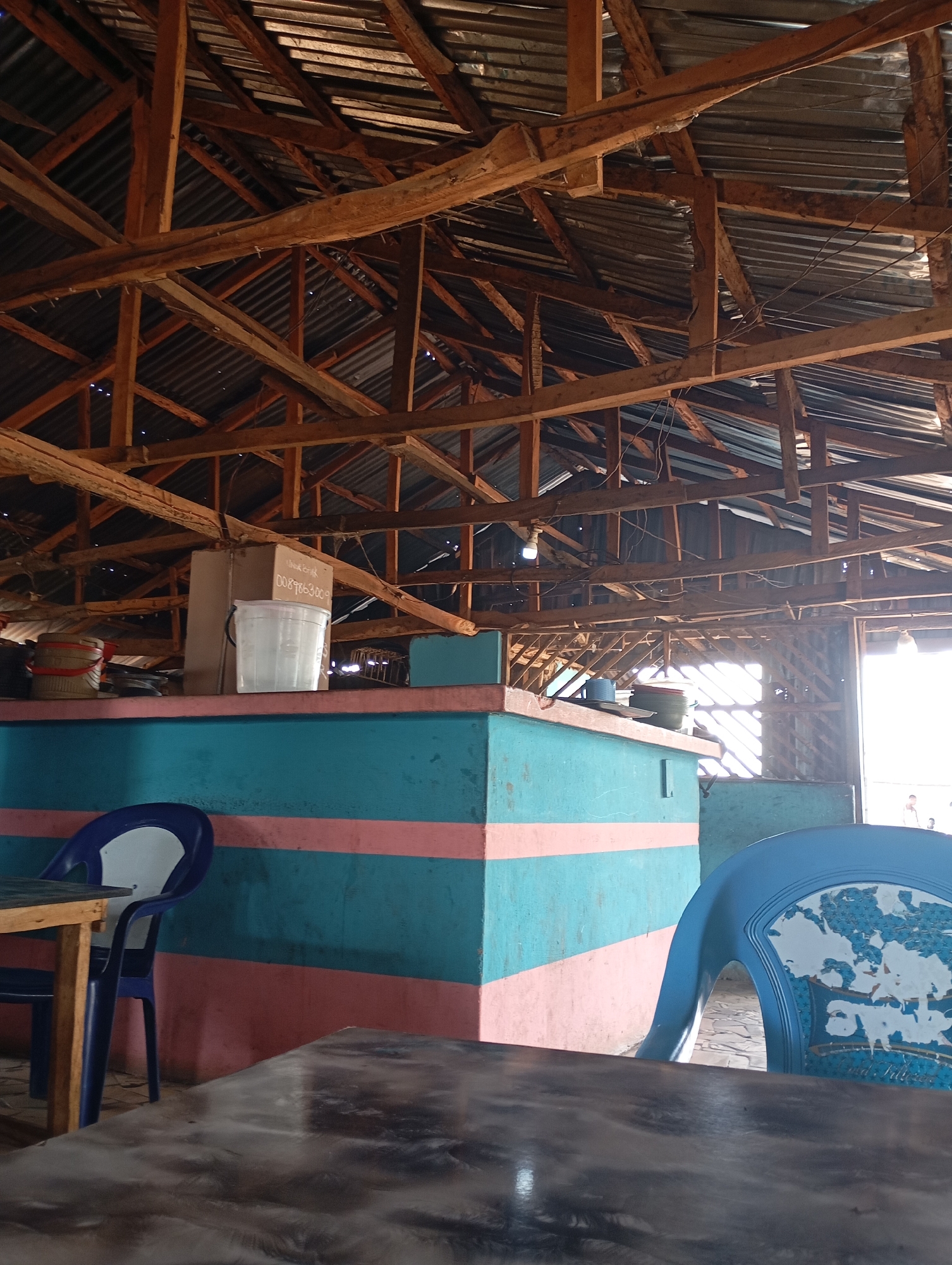 The image captures an interior view of a simple, open-air establishment with a corrugated metal roof supported by a network of wooden beams and rafters. The walls are constructed with a combination of painted surfaces and what appears to be partial open-air or lattice-like walls, allowing natural light to enter.
In the foreground, two tables are visible. The closer table has a dark, reflective surface. To the left, a wooden table with a light wooden top stands next to a blue chair with a white seat. Further to the right, a portion of another blue chair with a patterned white backrest is visible.
A counter area is situated behind the tables. The counter is painted with a prominent turquoise color, accented with horizontal pink stripes. On the counter, a white plastic bucket filled with water sits next to a cardboard box. Several other containers and items are also present on the counter.
The overall scene suggests a casual, possibly rural, establishment. The setting appears to be a local eatery or shop. The light suggests it is daytime. The atmosphere is utilitarian and functional. The image is taken from a seated perspective, looking towards the counter area and the structure of the building.
