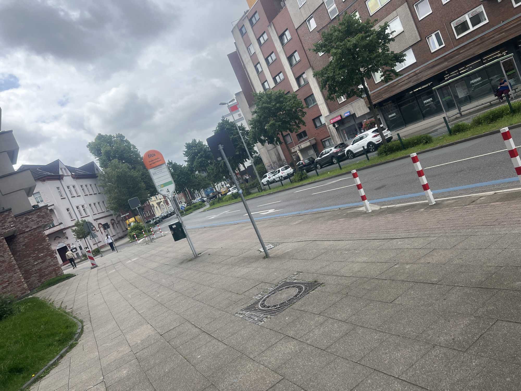 This image depicts an urban street scene in Herne, Germany, under a cloudy sky. The photograph is taken from a slightly low, angled perspective, looking across a wide, grey paved sidewalk towards a road and buildings.
In the immediate foreground, the concrete sidewalk shows a distinct rectangular manhole cover with a decorative circular grate. Red and white striped poles line the curb, separating the sidewalk from the asphalt road, which features a prominent blue line painted along its length.
Further along the sidewalk, a bus stop is visible, marked by an orange sign. The sign displays German text including "Herner Stadtwerke", "H Haltestelle vorübergehend aufgehoben" (bus stop temporarily suspended), "303 Röhling Hs.", and the route number "4". A transparent bus shelter is present, with a person sitting inside.
The road itself has several parked cars, mainly white and dark-colored sedans or hatchbacks. Across the street, multi-story residential or commercial buildings dominate the background. Some buildings feature red brick facades, while others have lighter colored panels. Green trees line the street, partially obscuring the lower floors of the buildings. Storefronts are visible on the ground floors of some buildings, with clear signage for "PENNY" (a supermarket), "PENNY Markt", and "DiSo Reisen". Another sign indicates "HERNER Stadtwerke". A few people are seen walking on the sidewalk in the distance. The scene suggests an ordinary daytime moment in a European town.