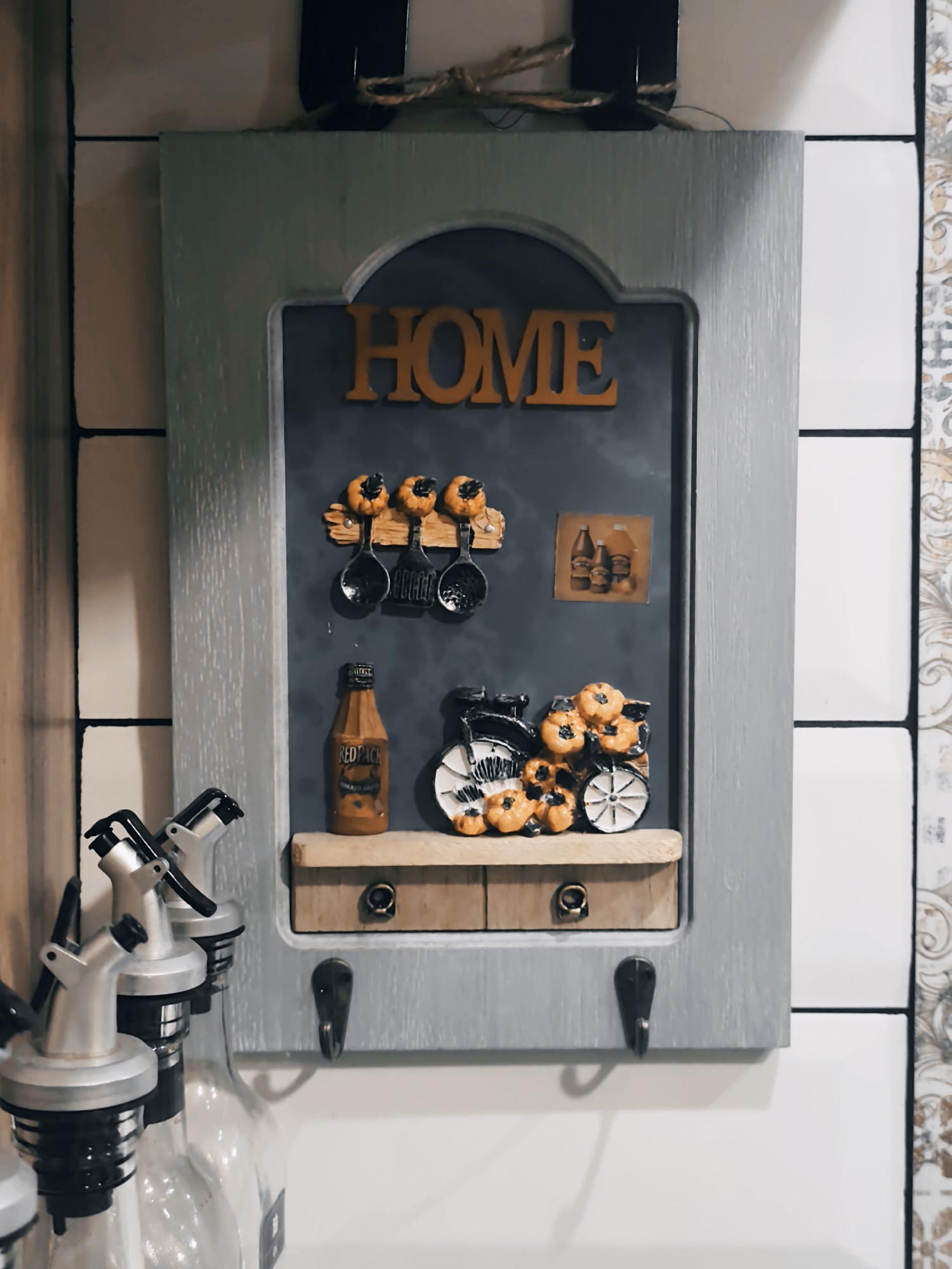 The image shows a decorative wall-mounted key holder with a distressed gray finish. It features an arched top and a dark gray backing board. The word "HOME" is spelled out in orange, raised lettering at the top. Below the word, there are three decorative spoons hanging from wooden pegs, each topped with a small, orange pumpkin. To the right of the spoons is a small, square picture depicting three bottles. Further down, a small wooden shelf holds a miniature bottle of "RED PACK Tomato Sauce" and a decorative item resembling a bicycle adorned with pumpkins. The shelf has two small, drawer-like fronts with circular knobs. Two black hooks are mounted on the bottom edge of the key holder. To the left of the key holder, partially visible, are the tops of three glass bottles with silver spouts, likely for oil or vinegar dispensers. The background consists of white tiled walls with thin dark grout lines, and a section of patterned wallpaper on the far right. The overall scene appears to be an indoor domestic setting, possibly a kitchen or entryway.