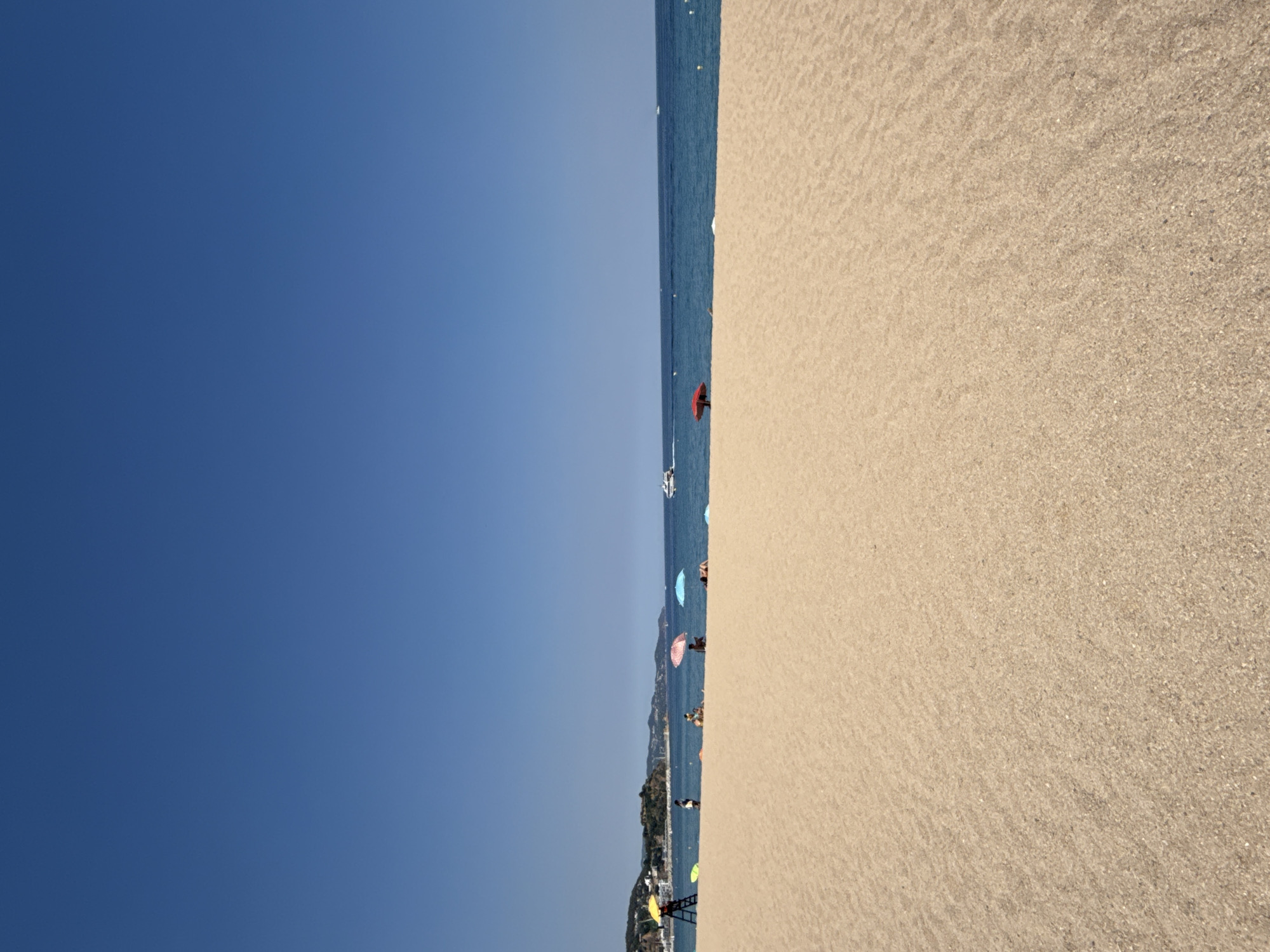 This media file depicts a sunny day at a beach in Blanes, Spain. The image is oriented vertically, with the sky occupying the left half and the beach and sea on the right. The sky is a clear, bright blue, indicating fair weather and likely daytime, possibly mid-morning or afternoon given the strong sunlight. The sea is a calm, deep blue, with a few small boats visible on the horizon.
The beach itself is covered in light-colored sand, with visible ripples suggesting the tide has recently receded or the wind is gently blowing. Scattered across the sand and near the water's edge are several colorful beach umbrellas, adding pops of red, pink, and blue to the scene. A few people are visible, some standing near the water, others appearing to be relaxing under the umbrellas. One person is clearly visible in the foreground, holding a red umbrella. The background shows a hilly, green coastline with buildings nestled along it, characteristic of a Mediterranean coastal town.
There is no visible text in the image. The overall impression is one of a pleasant, relaxing day at the beach, with the natural beauty of the coast and the sea complemented by the presence of beachgoers enjoying the sunny weather.