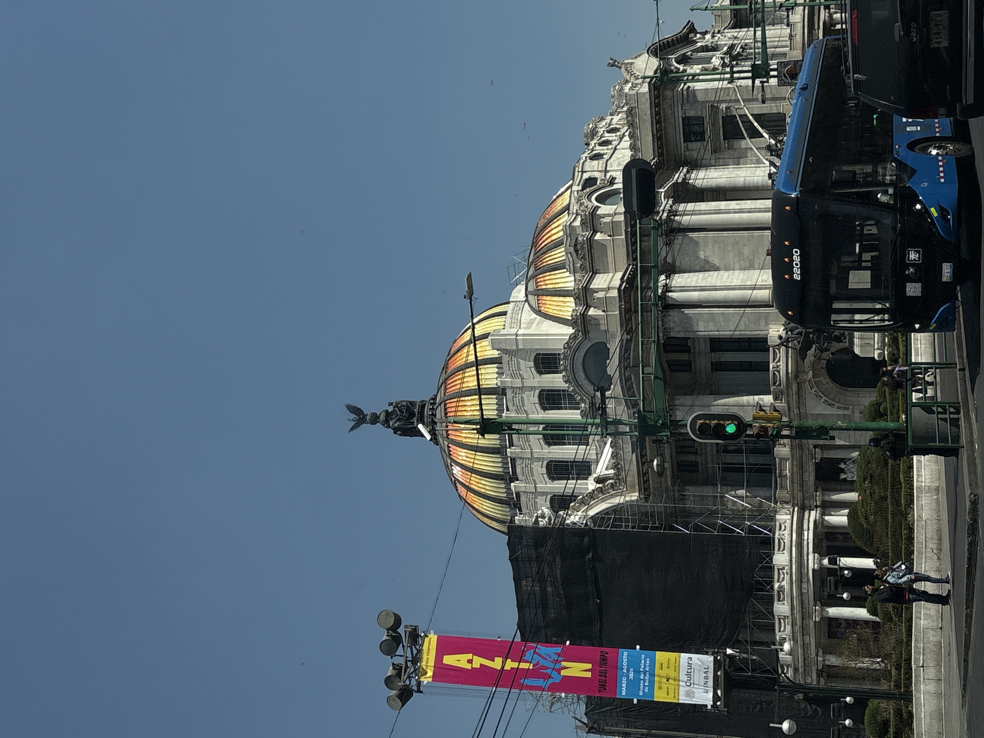 The image shows a street scene in Mexico City, Mexico. A grand, ornately decorated building with a large, striped dome dominates the background. A statue of an eagle tops the dome. In front of the building, there is a partially obscured construction area, covered with a black tarp and scaffolding. A tall banner advertising a temporary exhibition hangs near the construction, with the text "AZION TUNEL DEL TIEMPO MARZO-AGOSTO 2026 Museo del Palacio de Bellas Artes Cultura INBAL". A traffic light is positioned near the building, showing a green light. A bus is partially visible on the right side of the frame. Two pedestrians are walking on the sidewalk.