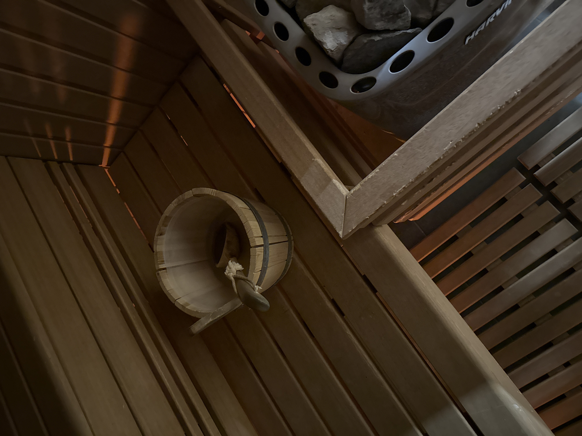 The image captures the interior of a sauna, possibly in a private residence, but located in Novi Sad, Serbia cannot be confirmed from the image alone. A wooden bucket and ladle are visible, positioned against the wooden wall of the sauna. Above, a sauna heater containing rocks, identified by the "Harvia" branding, is installed. The environment consists of wooden planks forming the walls and seating within the sauna. The lighting is subdued, typical of sauna environments, highlighting the wood grain and contributing to the ambiance.