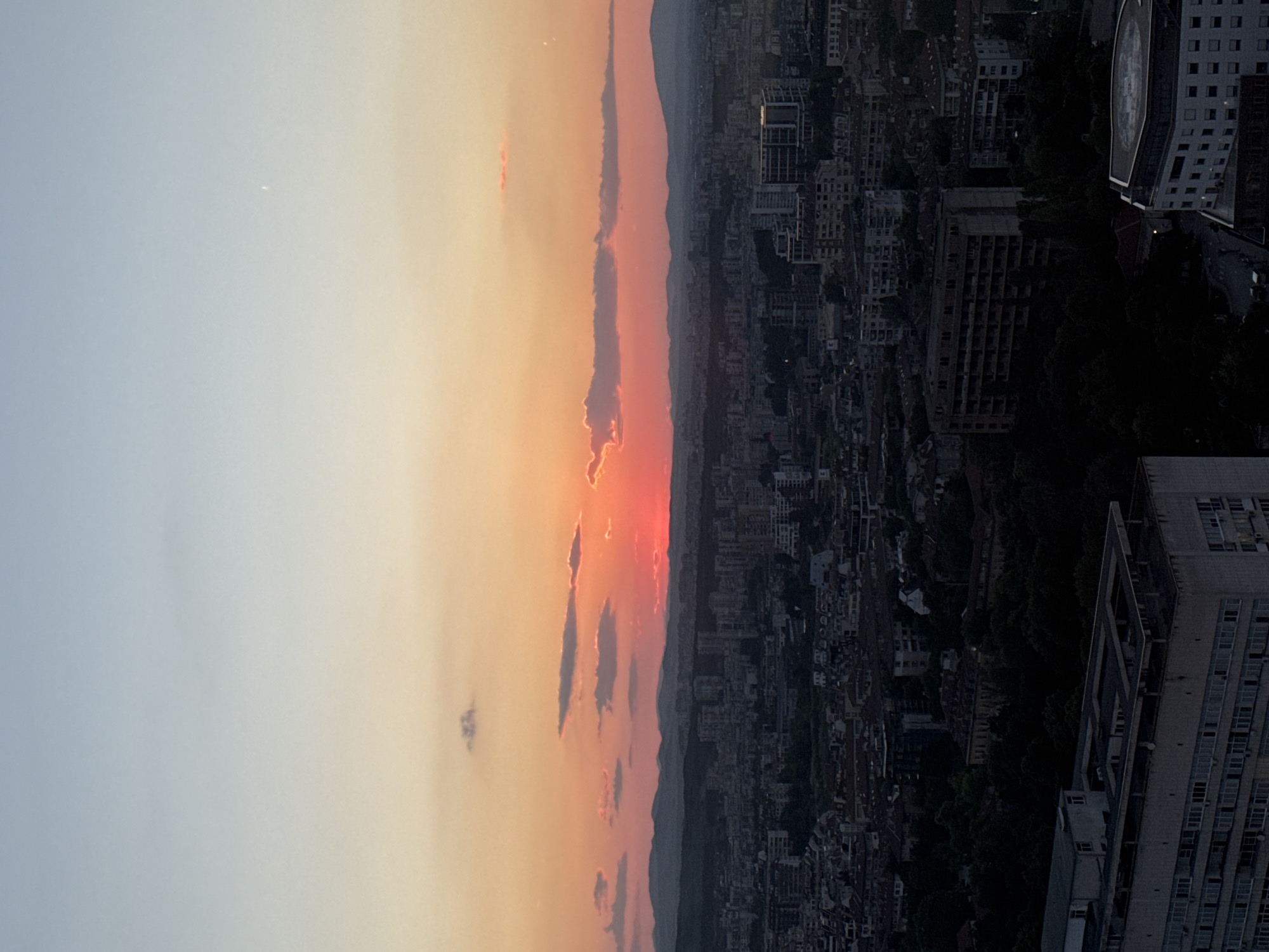 An aerial, wide-angle shot captures a cityscape at dusk. The sky transitions from a pale blue at the top to a gradient of orange and pink near the horizon, with the sun emitting a bright red glow just above a silhouetted mountain range. Several wispy clouds are scattered across the sky. The city below is densely populated with buildings of varying heights and architectural styles, appearing dark and somewhat indistinct due to the fading light. In the foreground, larger, more modern buildings with visible windows are prominent, contrasting with the older, lower-rise structures. Lush green trees are interspersed throughout the urban landscape, particularly in the midground and foreground. The overall scene depicts a large urban area under a dramatic sunset. The location is identified as Sofia, Bulgaria.