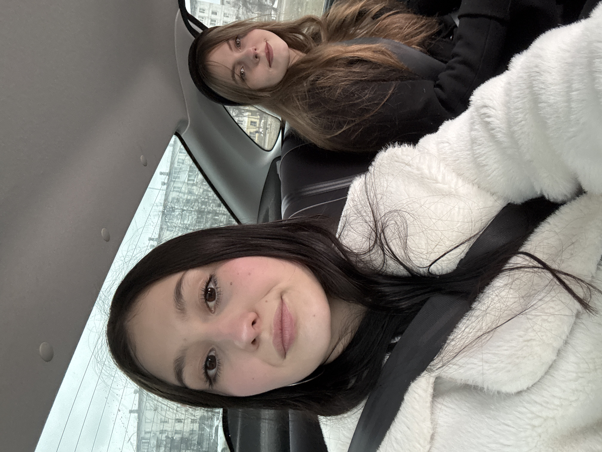 Two young women are sitting in a car. The woman in the foreground is wearing a white fuzzy jacket and a seatbelt. She has dark hair and is looking at the camera with a slight smile. The woman in the background has brown hair and a black headband, and she is also looking at the camera. Through the car window, buildings can be seen, suggesting an urban environment. The location is Chaikovsky, Russia.