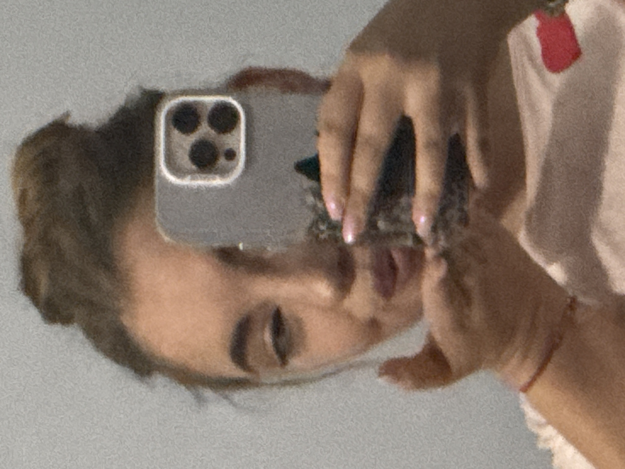 The scene depicts an individual taking a mirror selfie in an indoor environment in the city of Sofia, Bulgaria. A person with medium-brown hair styled in a high bun is visible, with their face partially obscured by a smartphone. The individual's left eye is closed or winking, and their lips are pursed in a gesture consistent with blowing a kiss. A light-colored smartphone, identified as an iPhone model with three dark camera lenses arranged in a square module, is held by the person's right hand. A thin, red string bracelet is present on the individual's left wrist, and their left hand is positioned near their mouth. The person is wearing a light-colored top featuring a small, bright red heart-shaped design on the lower right side. The background is a plain, light grey wall with a noticeable grainy texture.