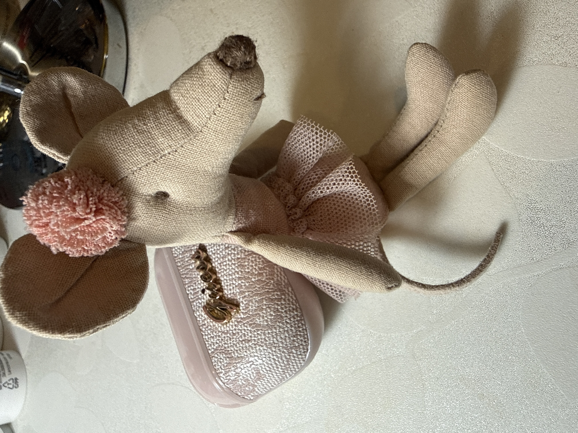 A stuffed toy mouse, dressed in a pink tutu, sits on a textured white surface. The mouse has large ears, one adorned with a pink pom-pom. A small, pink, textured case with a gold cursive "Guess" logo is positioned behind and to the left of the mouse. The background includes out-of-focus white containers and a reflective metallic object. The scene is indoors, on a tabletop. No specific location in Marum, Netherlands is visible.