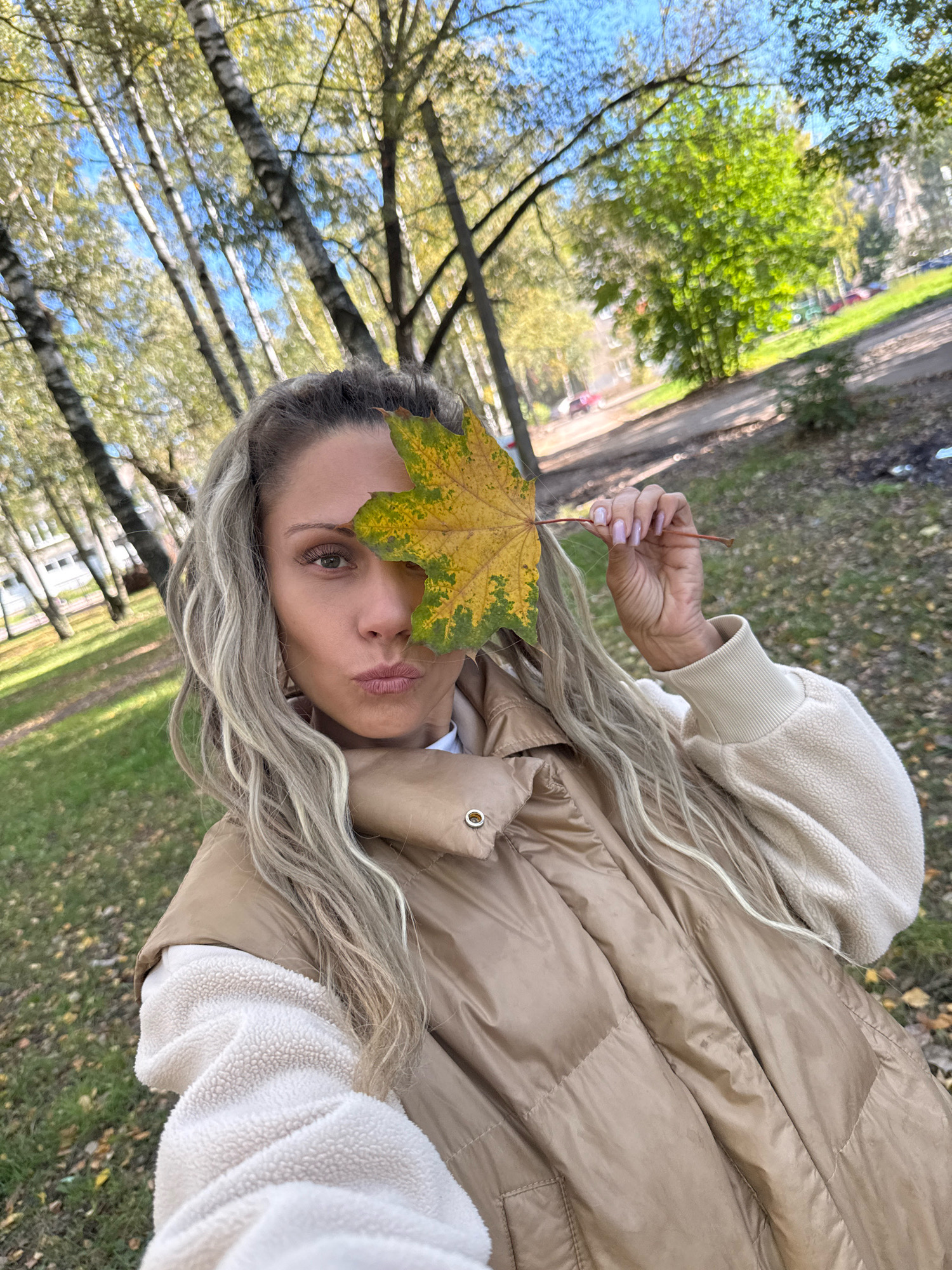 In Rusanovo, Russia, a woman with long blonde, wavy hair is pictured outdoors. She is wearing a tan puffer vest over a cream-colored fuzzy sweatshirt. She is holding a yellow and green maple leaf in front of her right eye and puckering her lips. The background shows a park with scattered autumn leaves on the ground and trees with dappled sunlight.