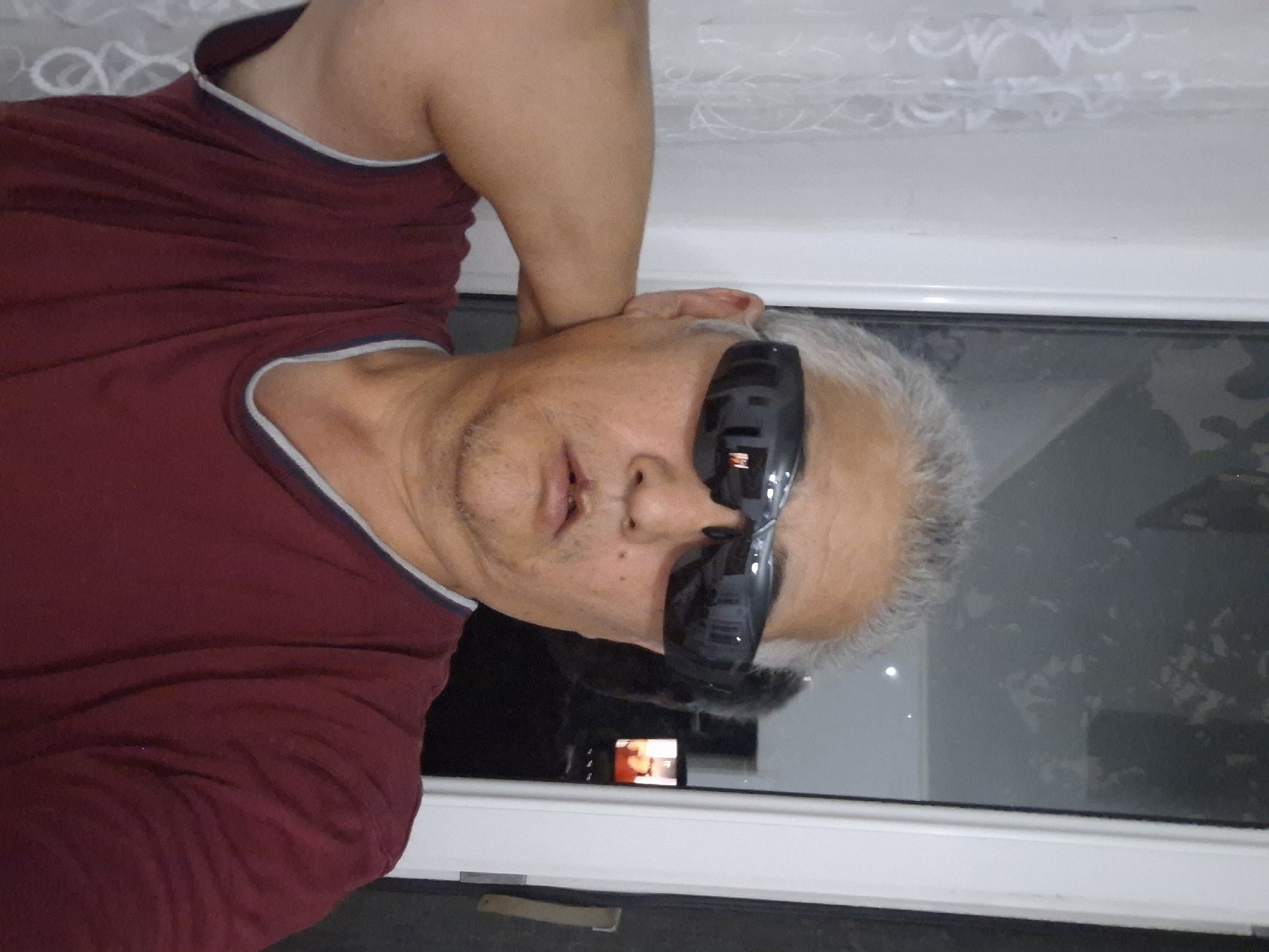 An individual with graying hair and sunglasses is posing for a selfie in what appears to be an indoor setting. The person is wearing a maroon t-shirt with a blue and gray trim around the collar. They are resting their left arm behind their head. In the background, a window reflects a dark environment with some indistinct shapes. To the right of the person, a door frame is visible. The overall environment suggests a residential or personal space. There is no visible information within the image that would indicate the specific location within Almaty, Kazakhstan.