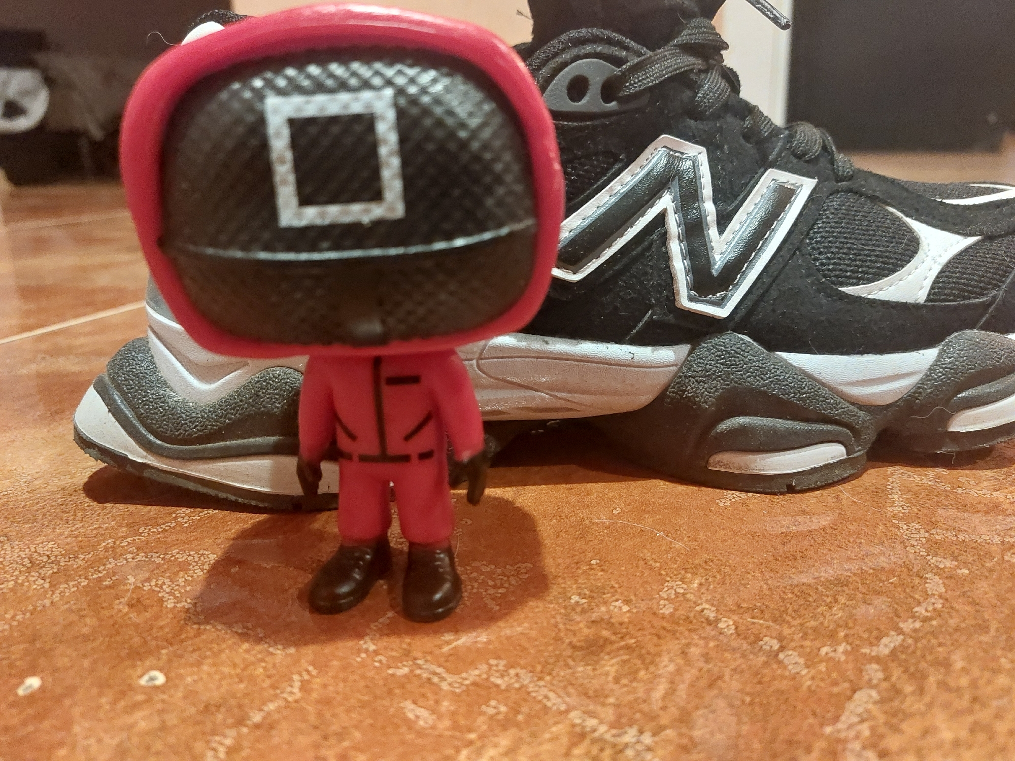 The image depicts a Funko Pop figure of a Squid Game guard with a square symbol on its mask, standing in front of a black and white New Balance sneaker. The background is blurred, but appears to be a brown tiled floor. There is no indication of the city of Tsarevo, Bulgaria in the scene. The figure is red, with a black mask and black shoes. The sneaker is black with white accents and the New Balance "N" logo. The lighting is diffused.