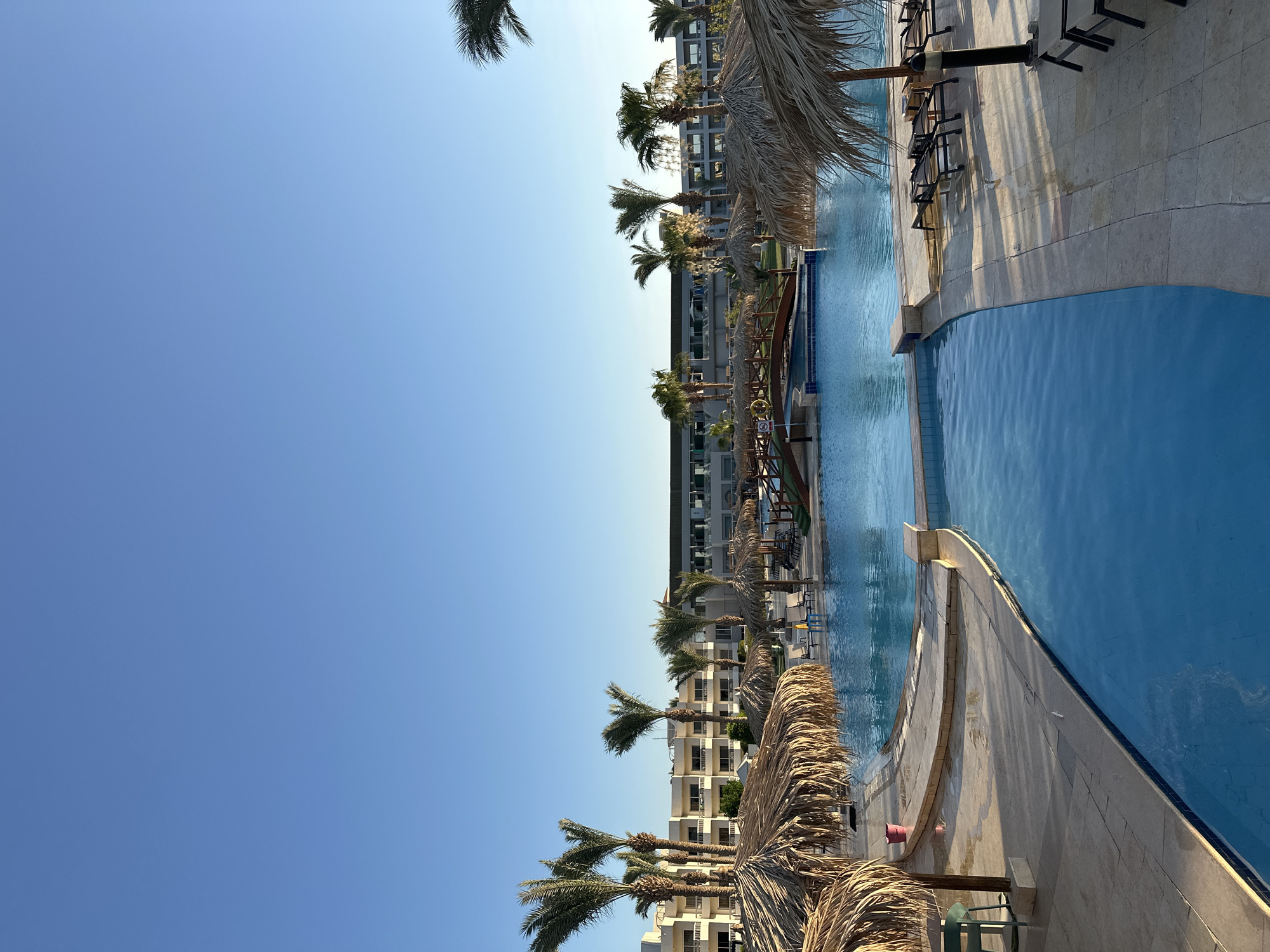 This image depicts a resort swimming pool area in Hurghada, Egypt, under a clear blue sky. The scene is dominated by a large, irregularly shaped swimming pool with bright blue water. A smaller, winding channel of water runs alongside the main pool.
Several thatched umbrellas and palm trees are scattered around the poolside, providing shade. Lounge chairs are visible on the tiled deck. In the background, a multi-story hotel building with many windows is situated behind a row of palm trees. A wooden bridge spans a section of the pool, and a small yellow life preserver hangs from its railing. The resort appears to be empty, with no people visible in the frame. The overall impression is one of a serene and well-maintained vacation destination.