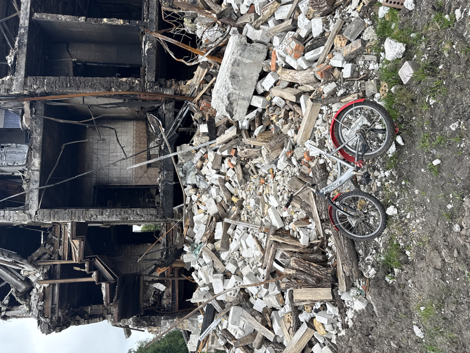 A dilapidated building stands in ruins. Its walls are charred and broken, with gaping holes where windows or doors once were. Piles of rubble and debris surround the building, consisting of bricks, concrete, and wooden planks. A child's bicycle lies on the ground, partially buried beneath the debris. The scene evokes a sense of destruction, loss, and the vulnerability of everyday objects amidst catastrophic events.